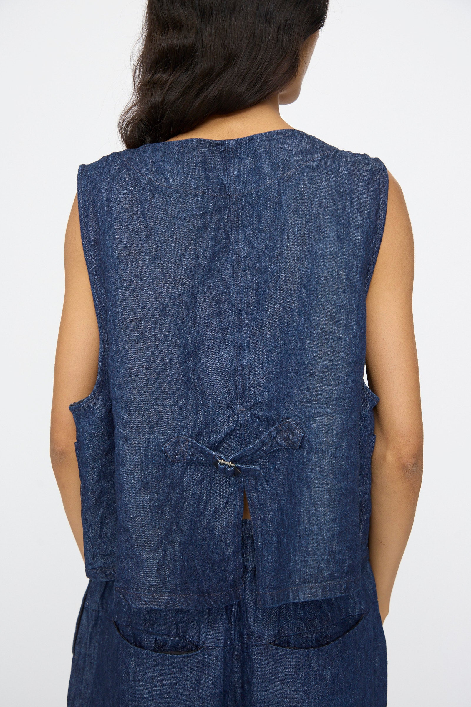 A person with long dark hair is shown from behind wearing the nest Robe 10oz. Linen Denim Vest in Indigo Blue, made in Japan, featuring a boxy fit, back tie detail, and matching bottoms.