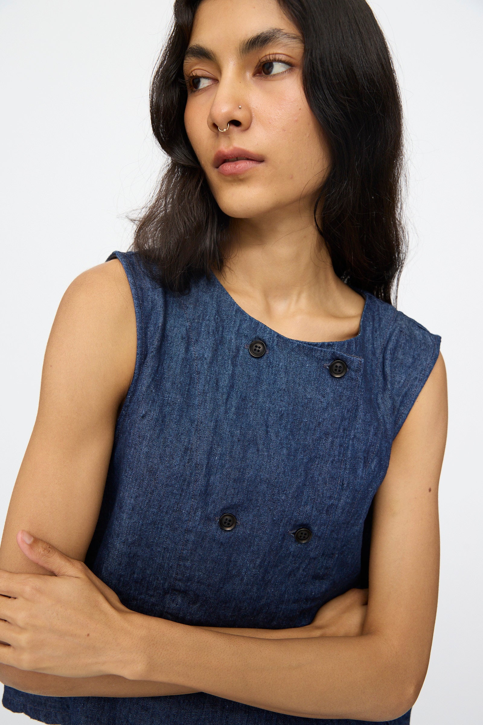 A person with long dark hair and a nose ring wears the nest Robe 10oz. Linen Denim Vest in Indigo Blue, made in Japan, standing with arms crossed and looking to the side.
