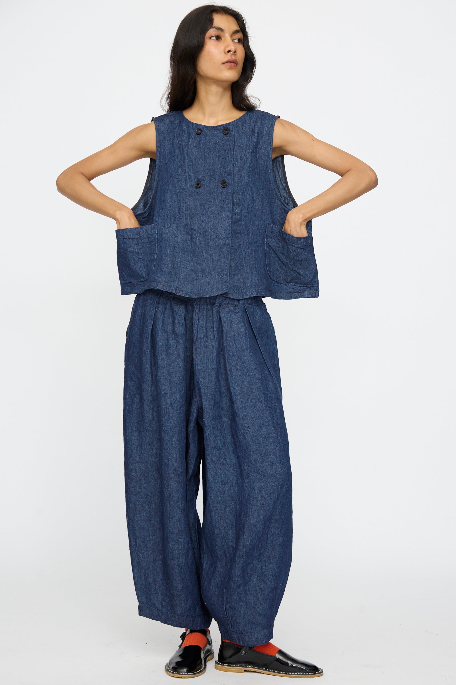 A person stands hands on hips against a plain background, wearing nest Robe’s 10oz. Linen Denim Vest in Indigo Blue, featuring a boxy, sleeveless design with large front pockets—crafted in Japan for a relaxed, stylish look.