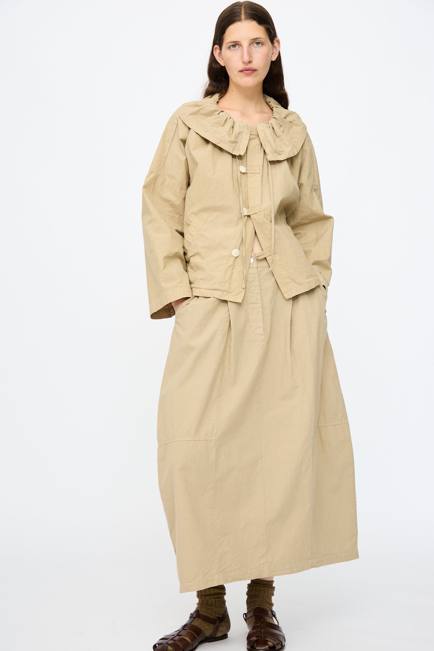 A woman wears the nest Robe Cotton Linen Dungaree Balloon Skirt in beige, paired with a beige long-sleeve buttoned jacket with a ruffled collar, brown sandals, and olive socks.