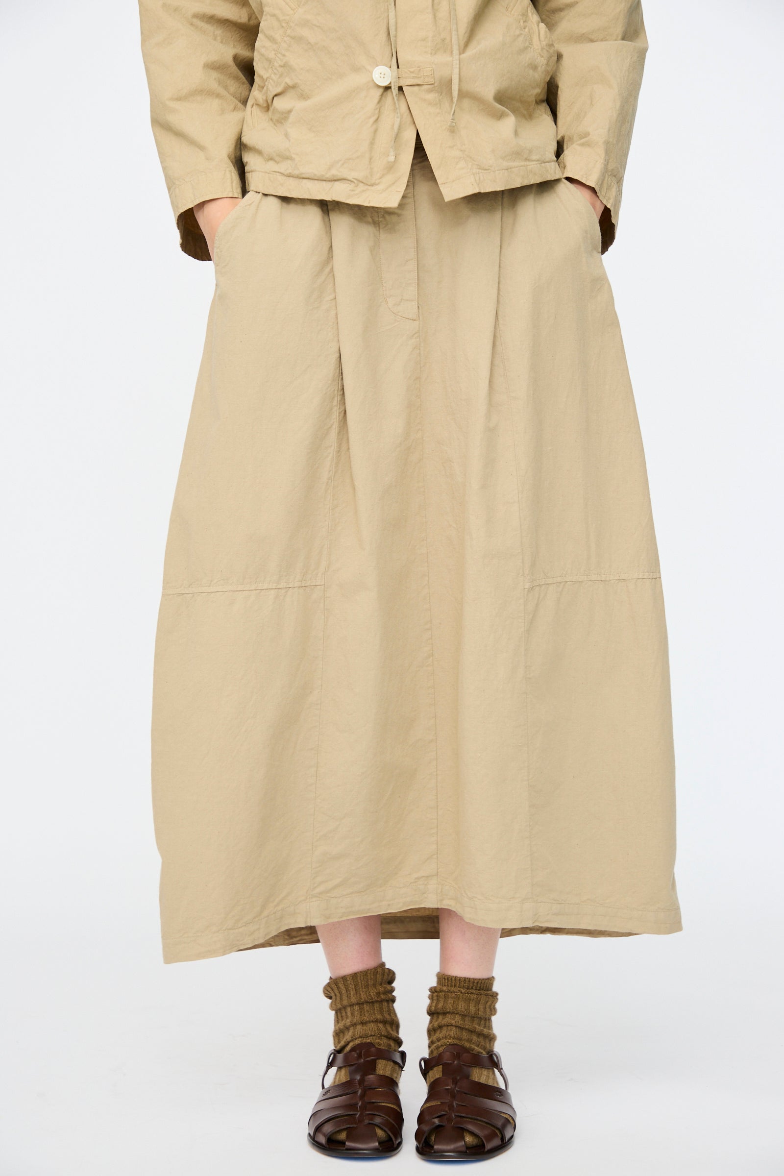 Model wears the nest Robe Cotton Linen Dungaree Balloon Skirt in Beige, styled with a matching beige jacket, brown socks, and brown sandals, standing with hands in pockets against a plain background.