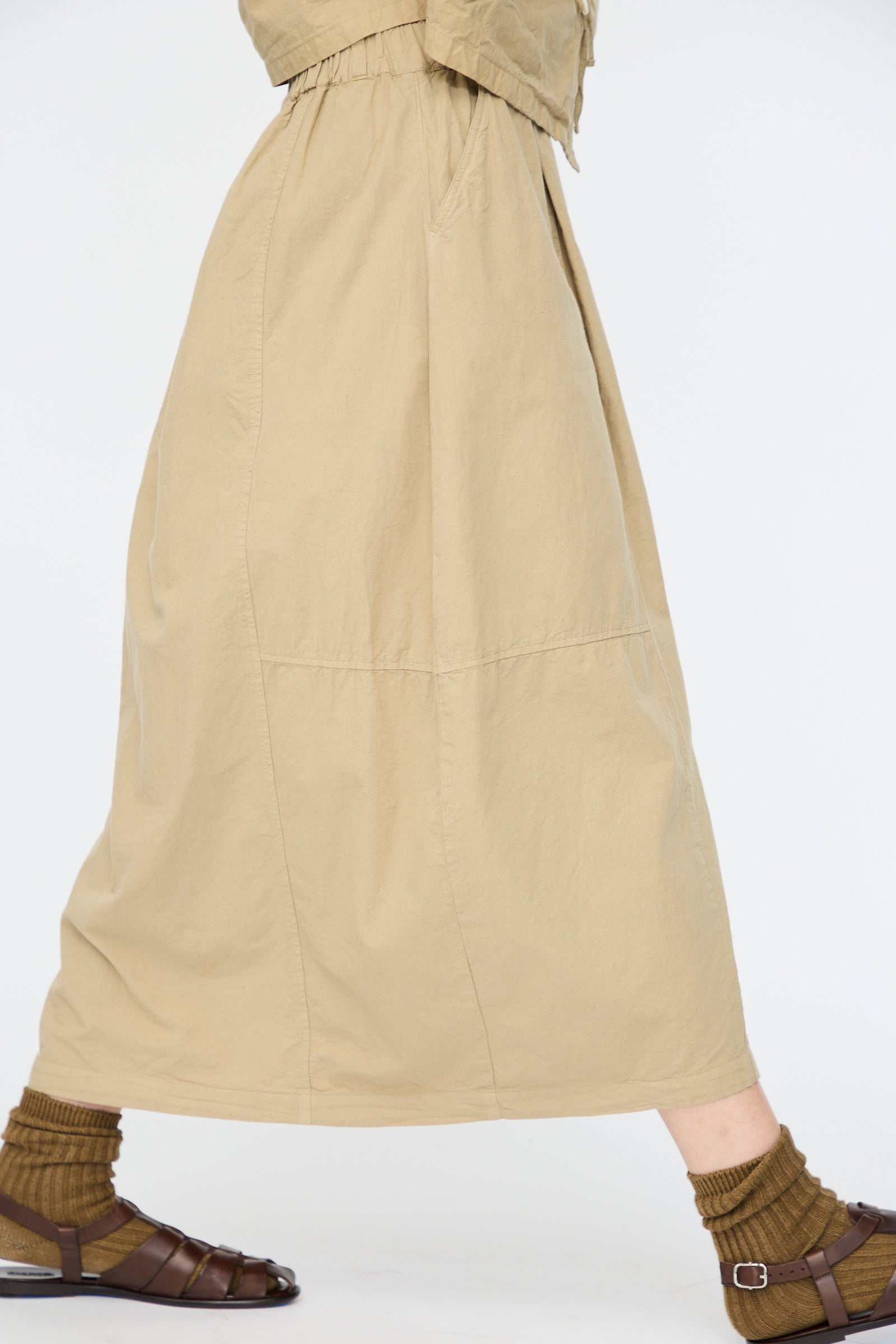 A person wearing nest Robe's Cotton Linen Dungaree Balloon Skirt in Beige, styled with brown socks and sandals, walks against a plain white background.