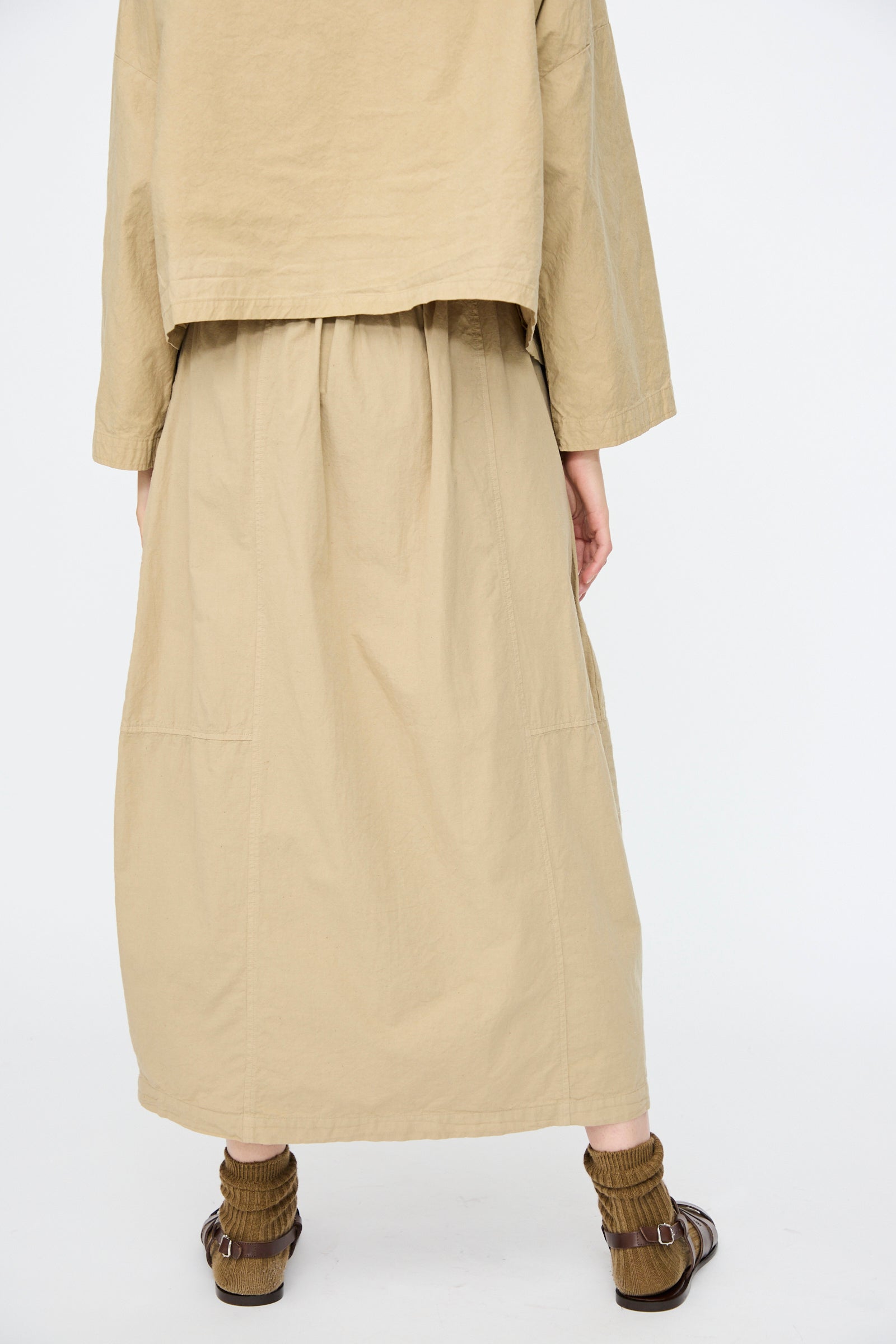 A person is seen from behind wearing the nest Robe Cotton Linen Dungaree Balloon Skirt in Beige with a loose tan long-sleeve top, brown socks, and open-toed sandals, set against a plain background.