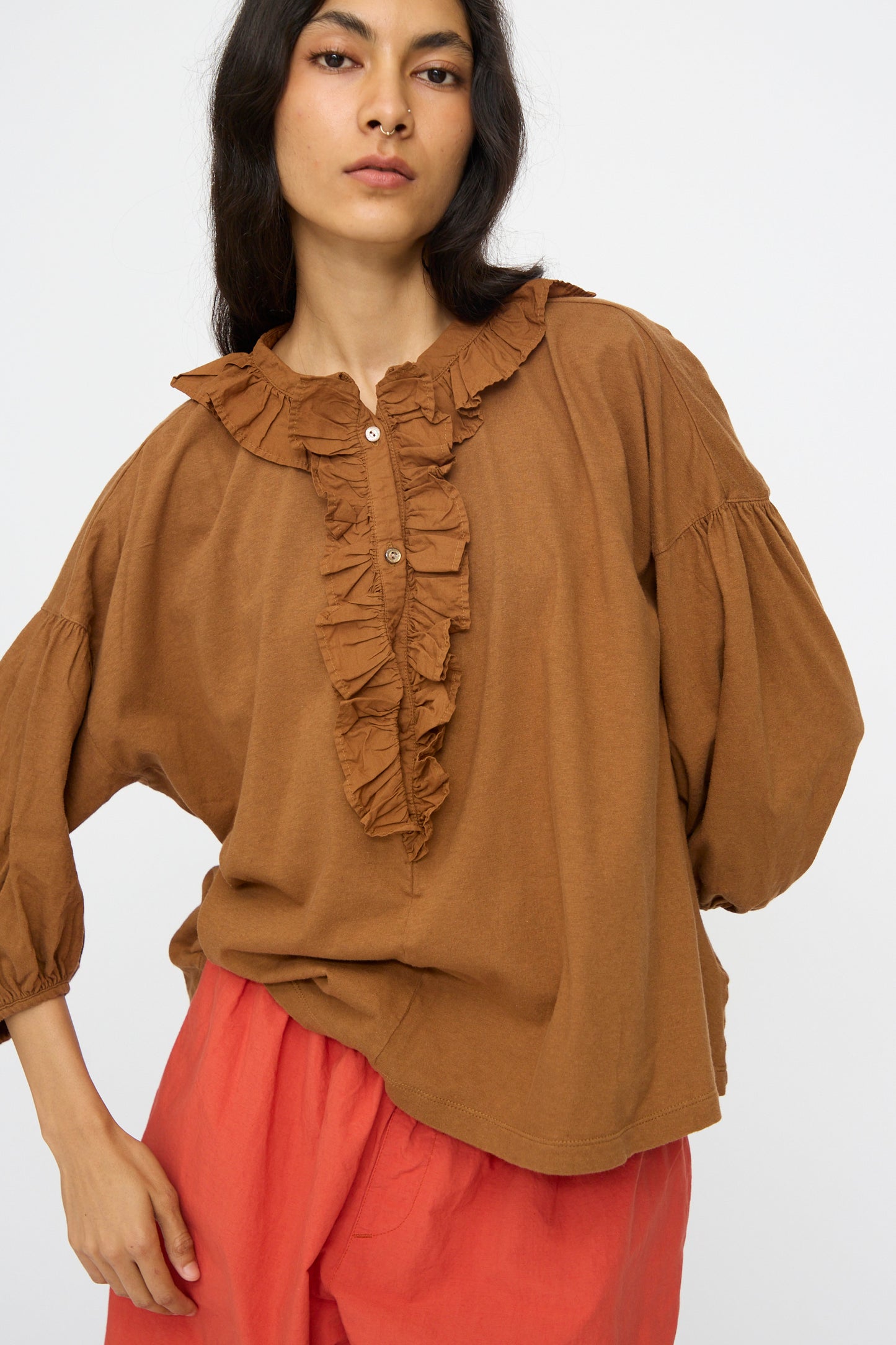 A woman wears the nest Robe Cotton Linen Frill Top in Brown with buttons, paired with coral pants, standing against a plain white background.