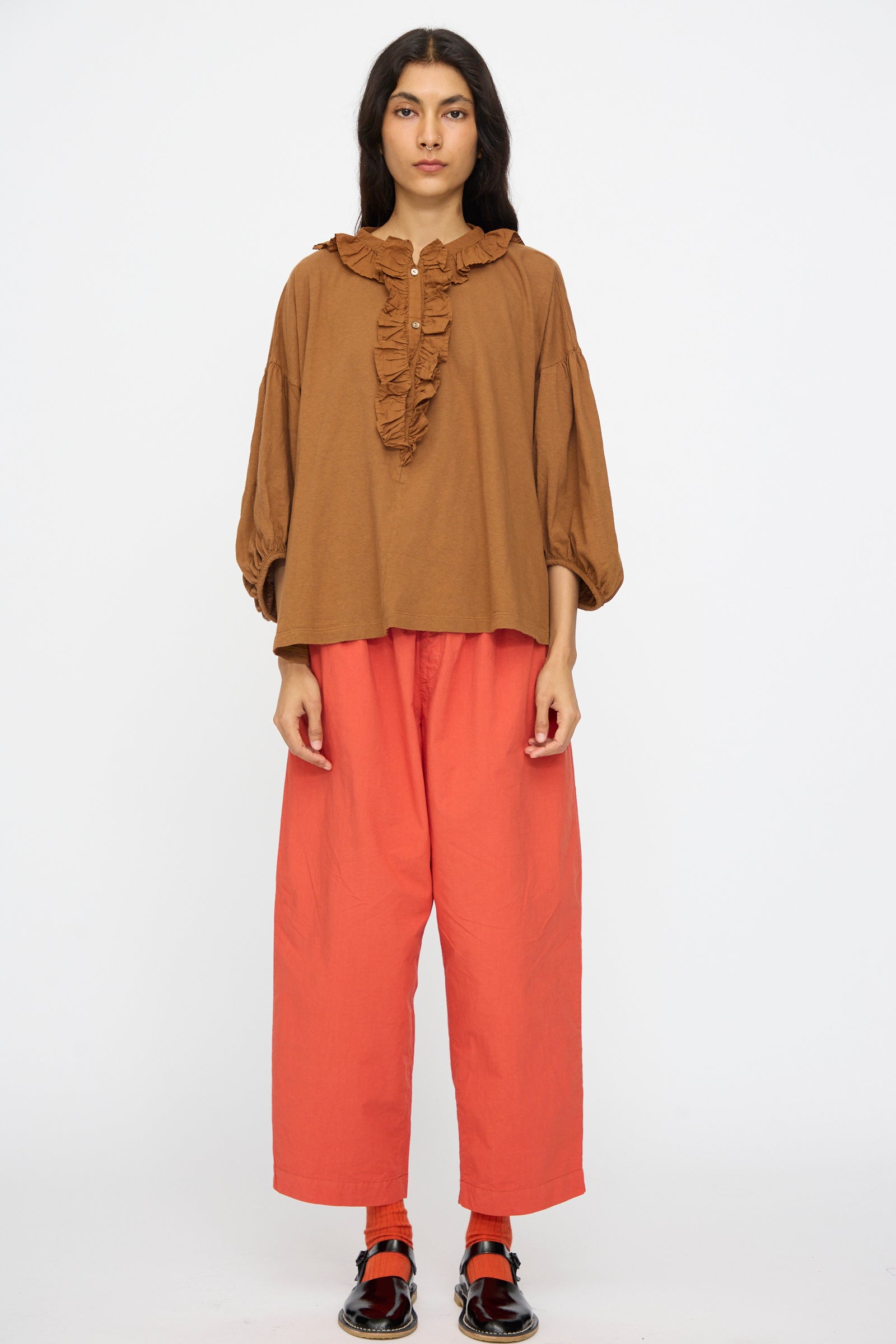 A person stands facing forward against a plain white background, wearing the nest Robe Cotton Linen Frill Top in Brown, wide red-orange pants, red socks, and black shoes.