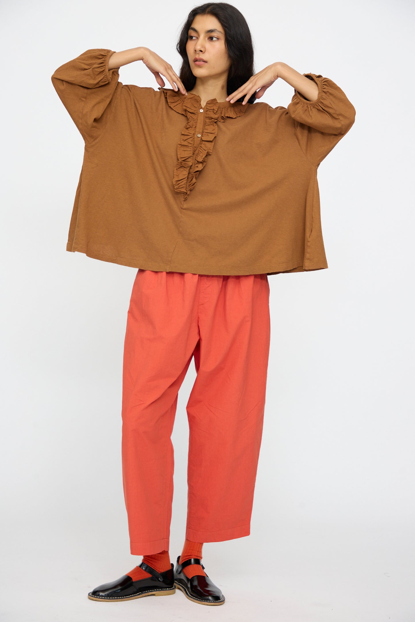 A person stands against a plain background wearing the nest Robe Cotton Linen Frill Top in Brown, red pants, red socks, and black shoes, with arms bent and hands near their shoulders.