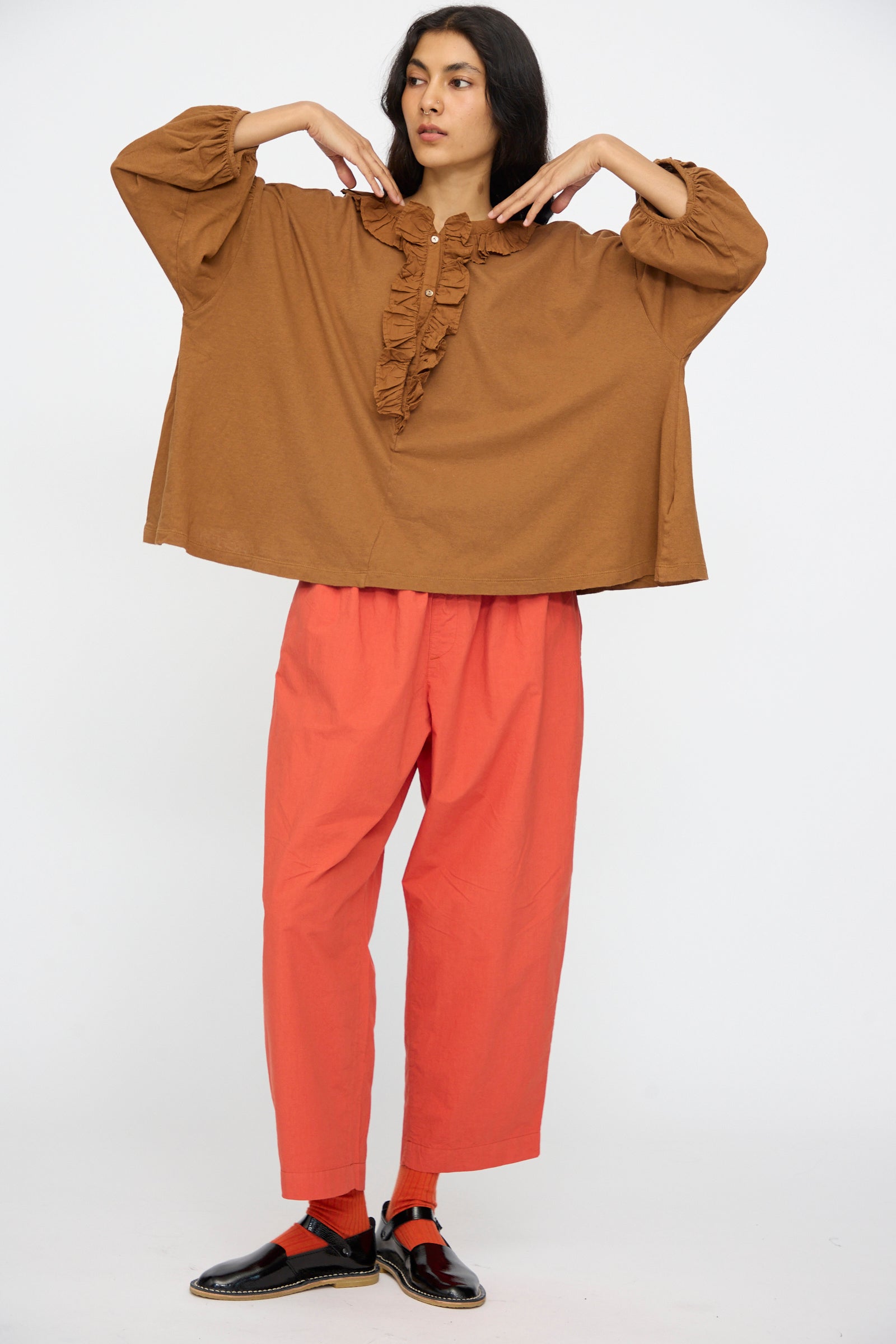 A person stands against a plain background wearing the nest Robe Cotton Linen Frill Top in Brown, red pants, red socks, and black shoes, with arms bent and hands near their shoulders.