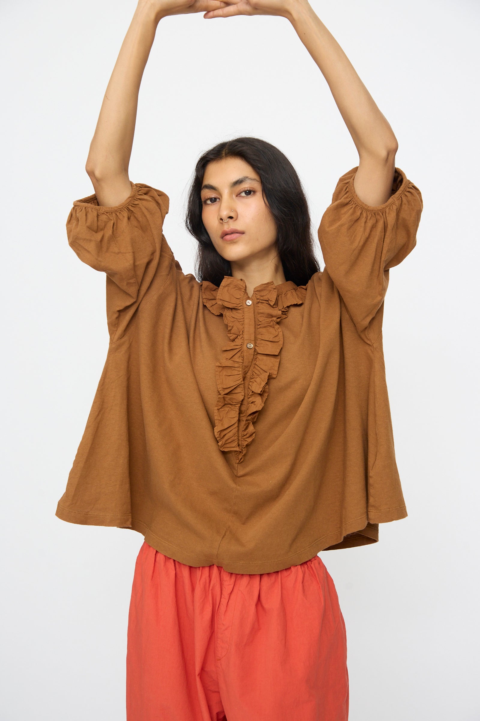 Model wears the nest Robe Cotton Linen Frill Top in Brown with red pants, standing with arms raised above head against a plain white background.