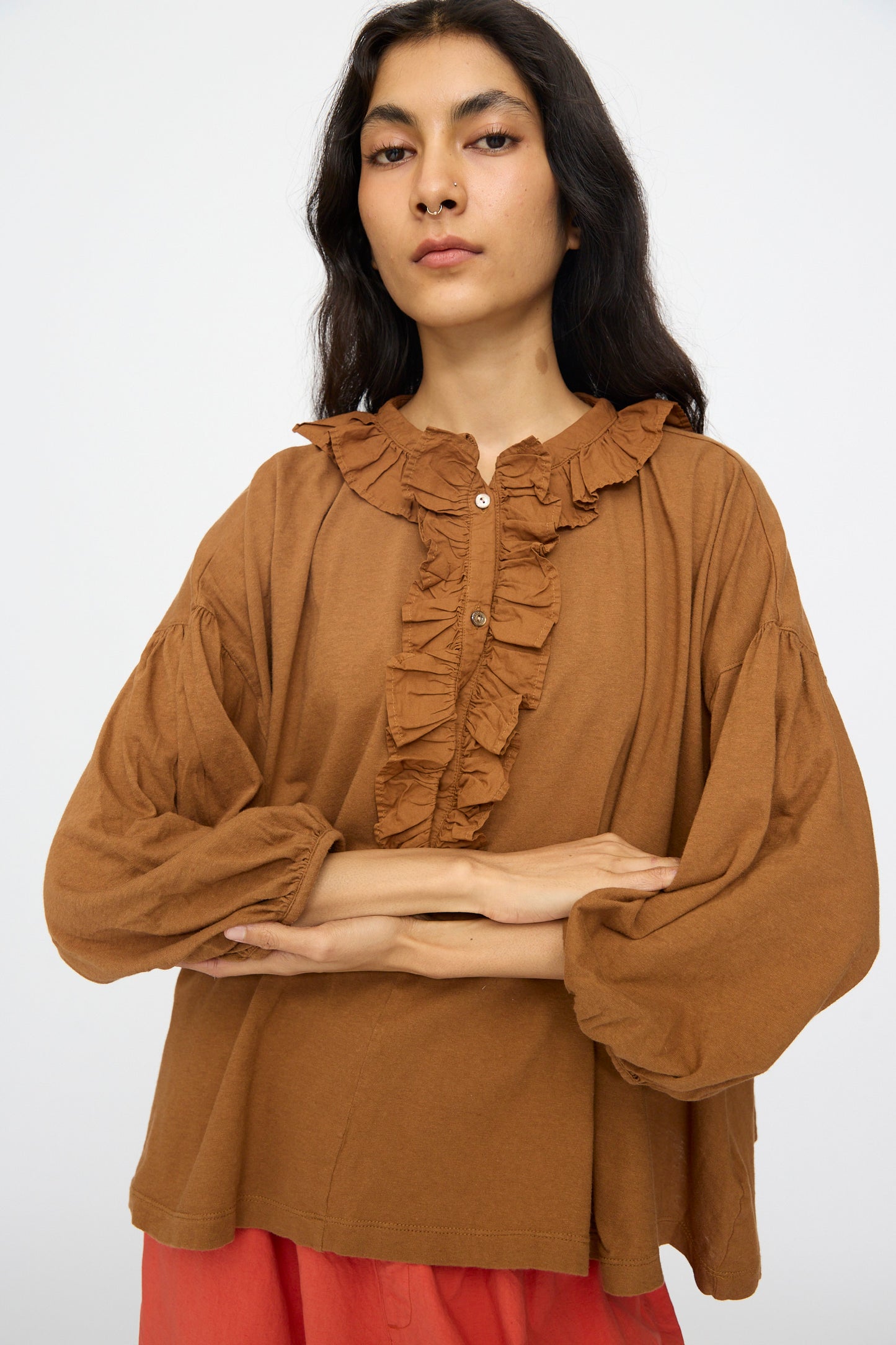 A woman with long dark hair wears the nest Robe Cotton Linen Frill Top in Brown, paired with red bottoms, standing against a plain light background with her arms crossed.