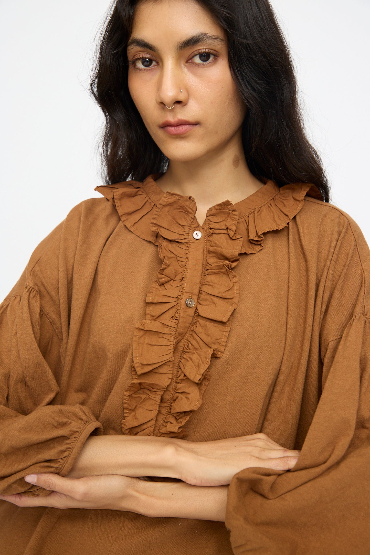 A person with long dark hair stands with arms crossed, wearing the nest Robe Cotton Linen Frill Top in Brown and looking directly at the camera.
