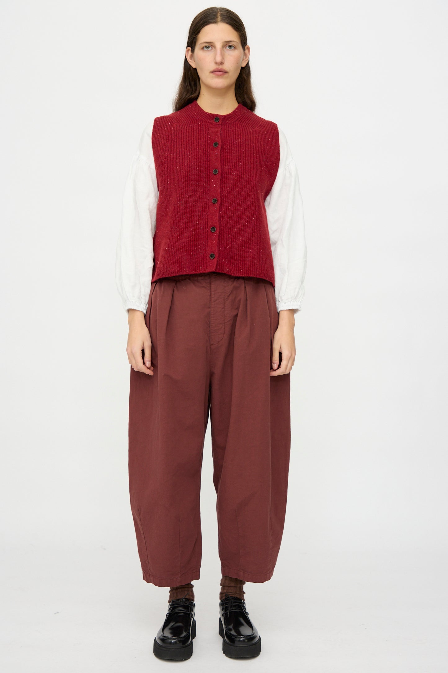 A person faces forward, wearing a red vest, white shirt, and nest Robe Cotton Linen Omi-Zarashi Pleated Pants in Brick, paired with black shoes against a plain white background.
