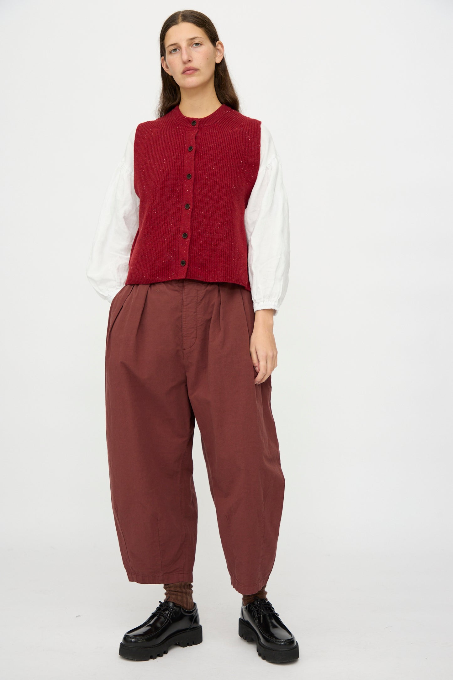A person stands against a plain background wearing a red vest, white long-sleeve shirt, and nest Robe’s Cotton Linen Omi-Zarashi Pleated Pants in Brick, made in Japan, paired with black platform shoes.