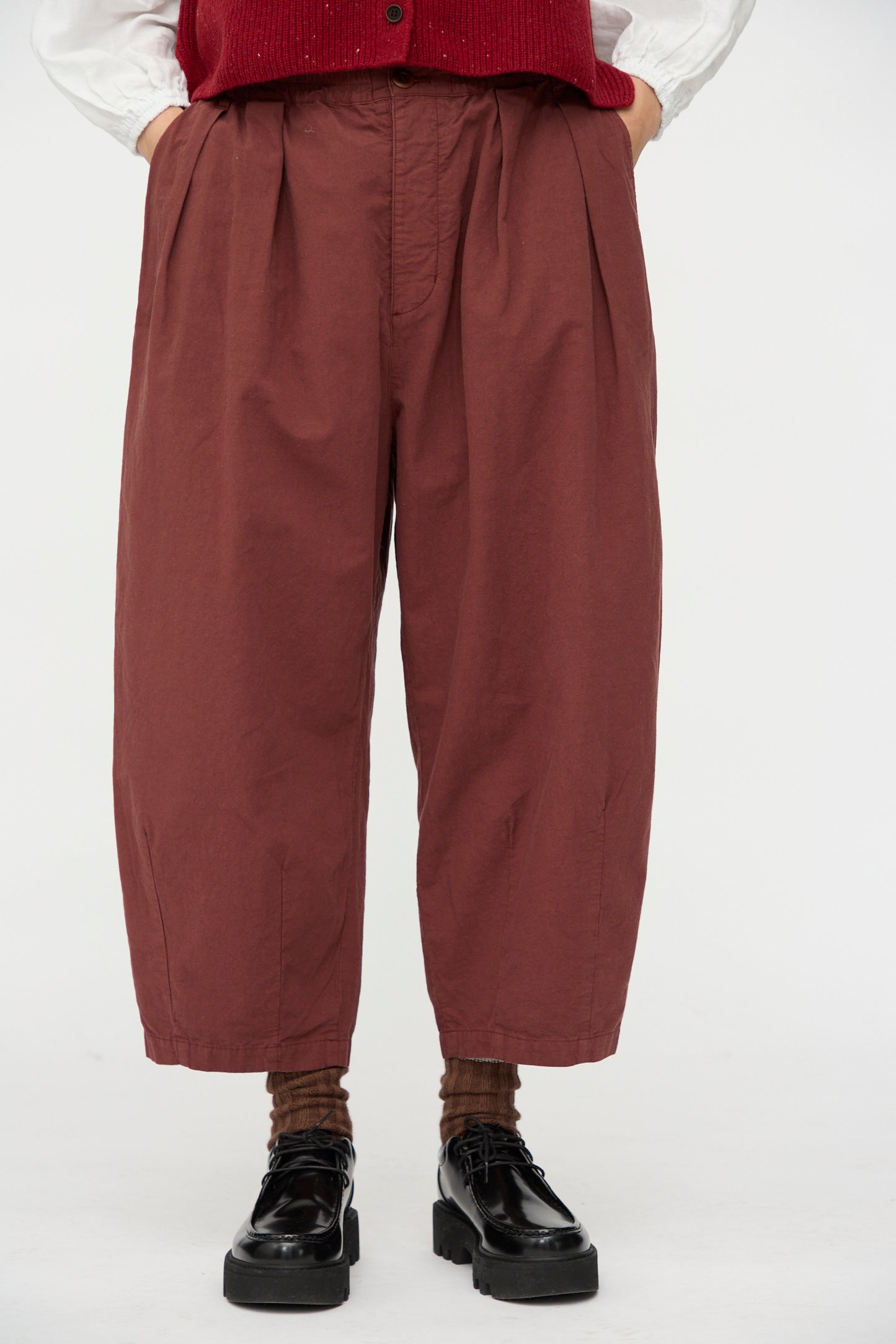 A person is wearing nest Robe’s Cotton Linen Omi-Zarashi Pleated Pants in Brick, made in Japan, paired with brown socks, black shoes, a white shirt, and a partially visible red vest.