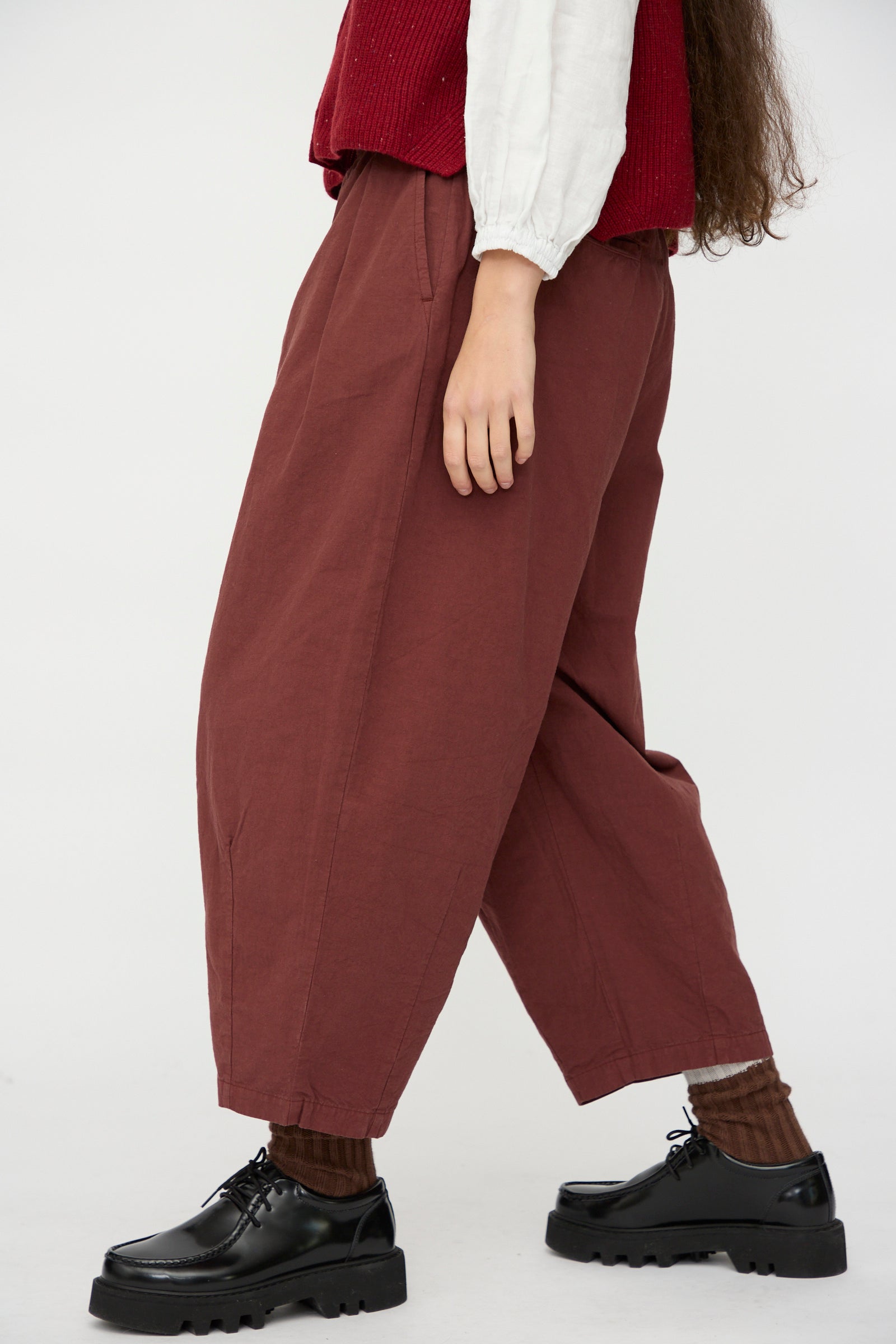 Person wearing nest Robe's Cotton Linen Omi-Zarashi Pleated Pants in Brick, a white long-sleeve shirt, red vest, brown socks, and black platform shoes stands against a plain white background. 