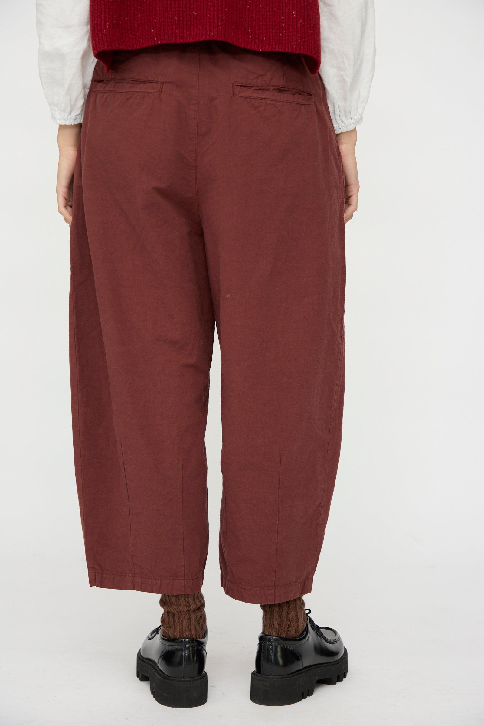 A person stands with their back to the camera, wearing nest Robe Cotton Linen Omi-Zarashi Pleated Pants in Brick, a white top, red vest, brown socks, and black shoes against a plain background.