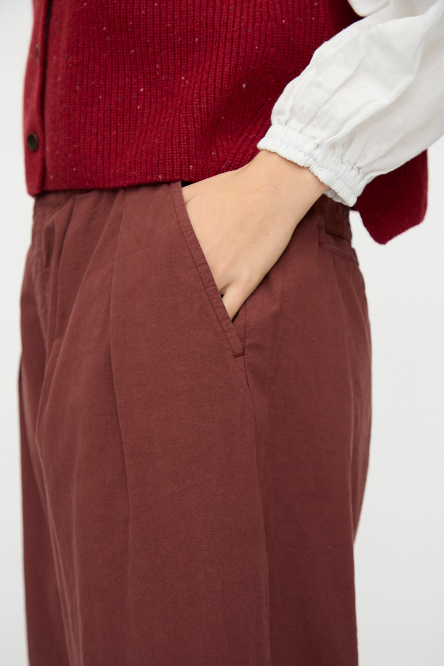 A person wearing nest Robe’s Cotton Linen Omi-Zarashi Pleated Pants in Brick, paired with a white long-sleeve shirt and red vest, has one hand in their pocket.