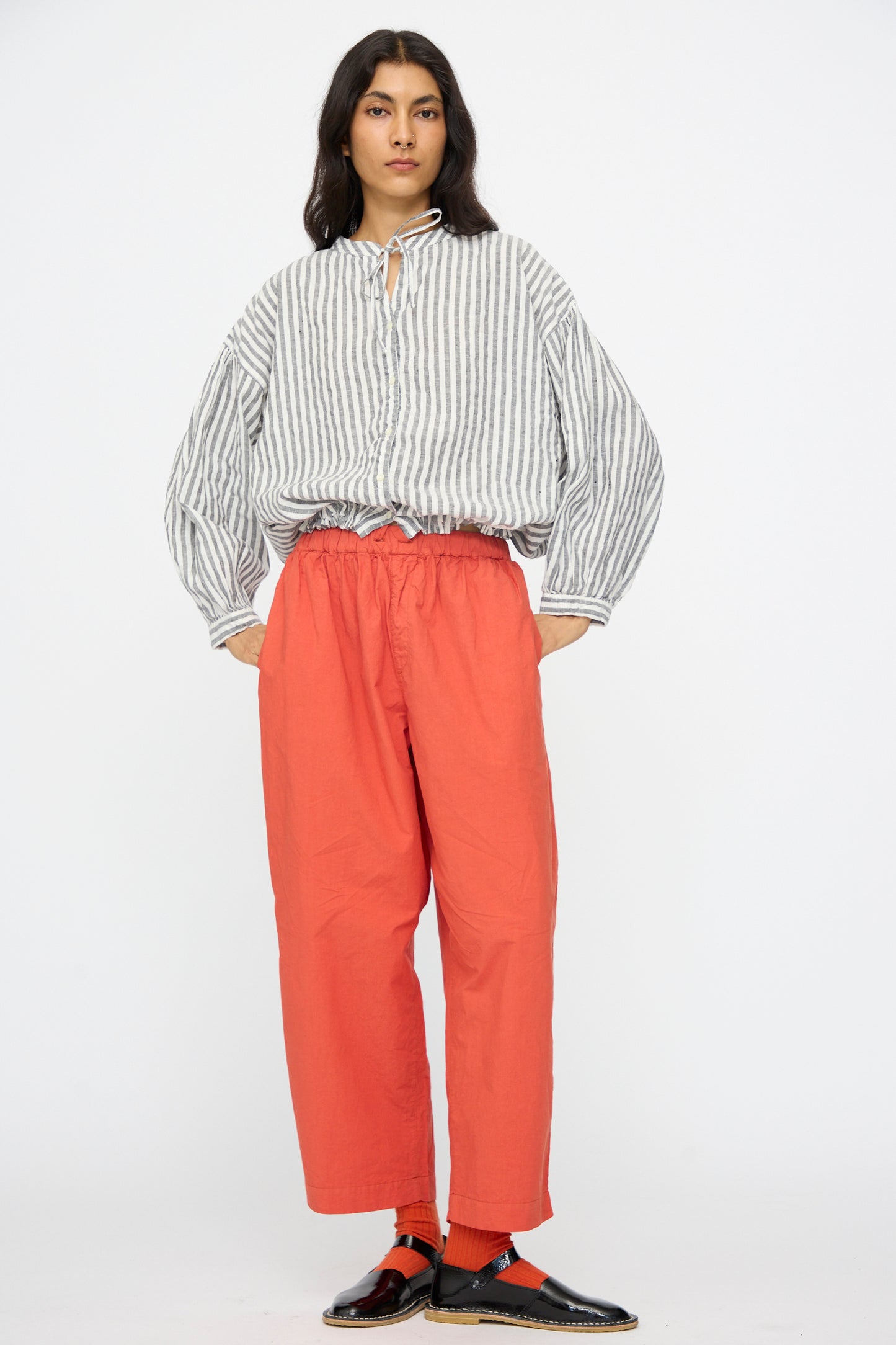A person stands against a white background wearing a gray and white striped blouse, orange Cotton Linen Typewriter Omi-Zarashi Easy Pants by nest Robe, orange socks, and black slip-on shoes.