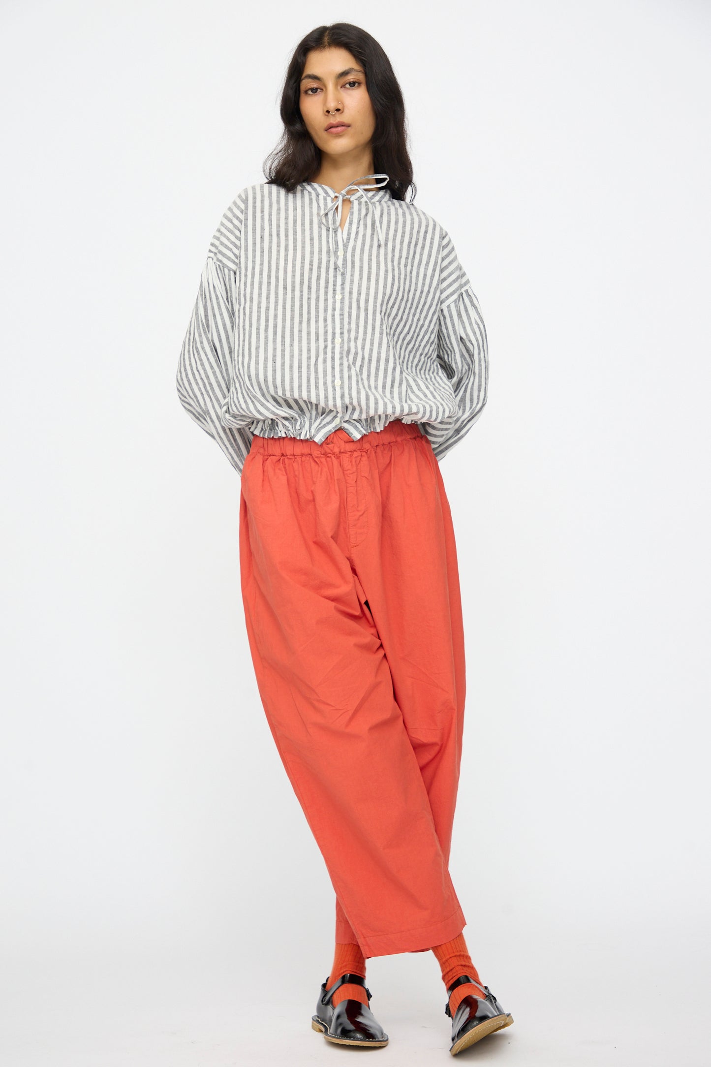 A person stands against a plain background wearing a loose, striped shirt and nest Robe’s Cotton Linen Typewriter Omi-Zarashi Easy Pants in Orange, paired with sandals and socks.