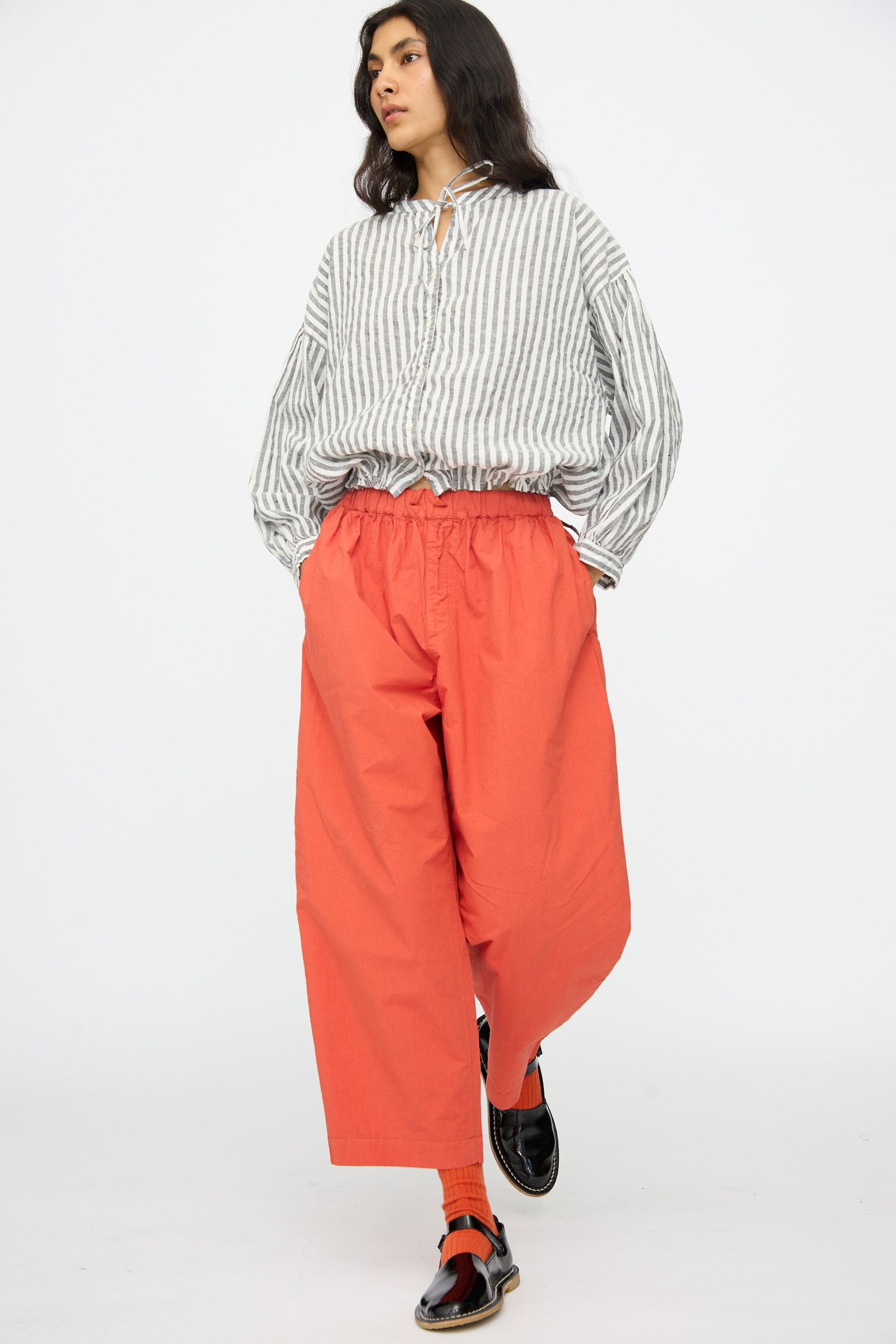 A person stands against a white background wearing nest Robe's Cotton Linen Typewriter Omi-Zarashi Easy Pants in Orange, a loose striped button-up shirt, black loafers, and orange socks.