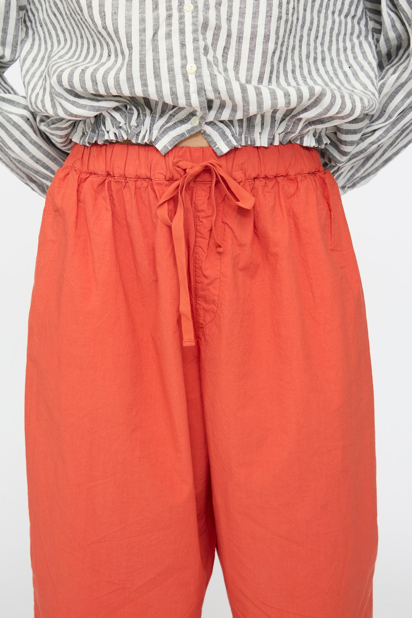 A person wears nest Robe’s Cotton Linen Typewriter Omi-Zarashi Easy Pants in Orange—high-waisted, wide-leg with a drawstring waist—paired with a gray and white striped button-up shirt tucked in.