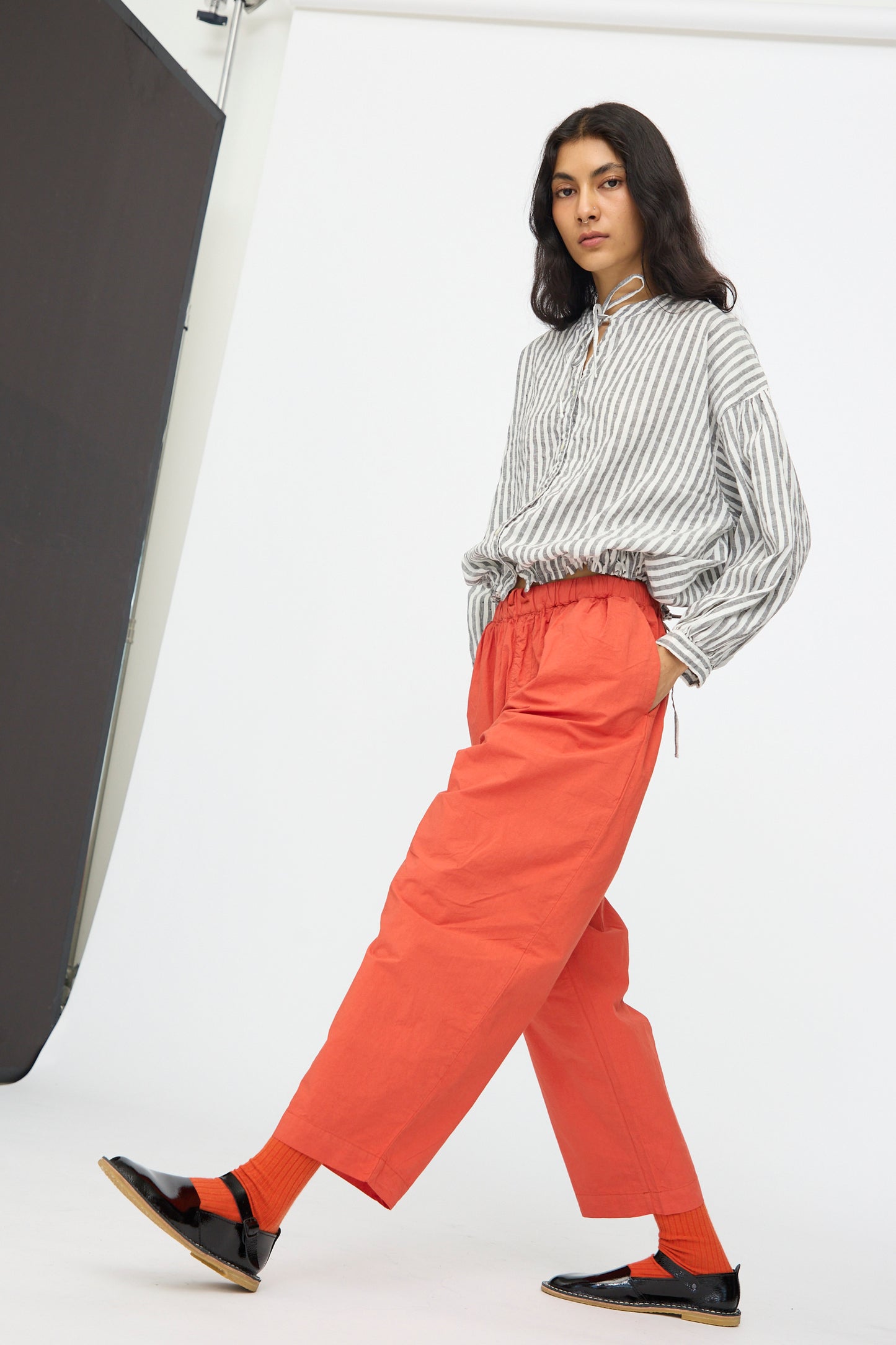 A person stands in a studio against a white background, wearing a gray and white striped shirt, orange socks, black shoes, and nest Robe’s Cotton Linen Typewriter Omi-Zarashi Easy Pants in Orange.