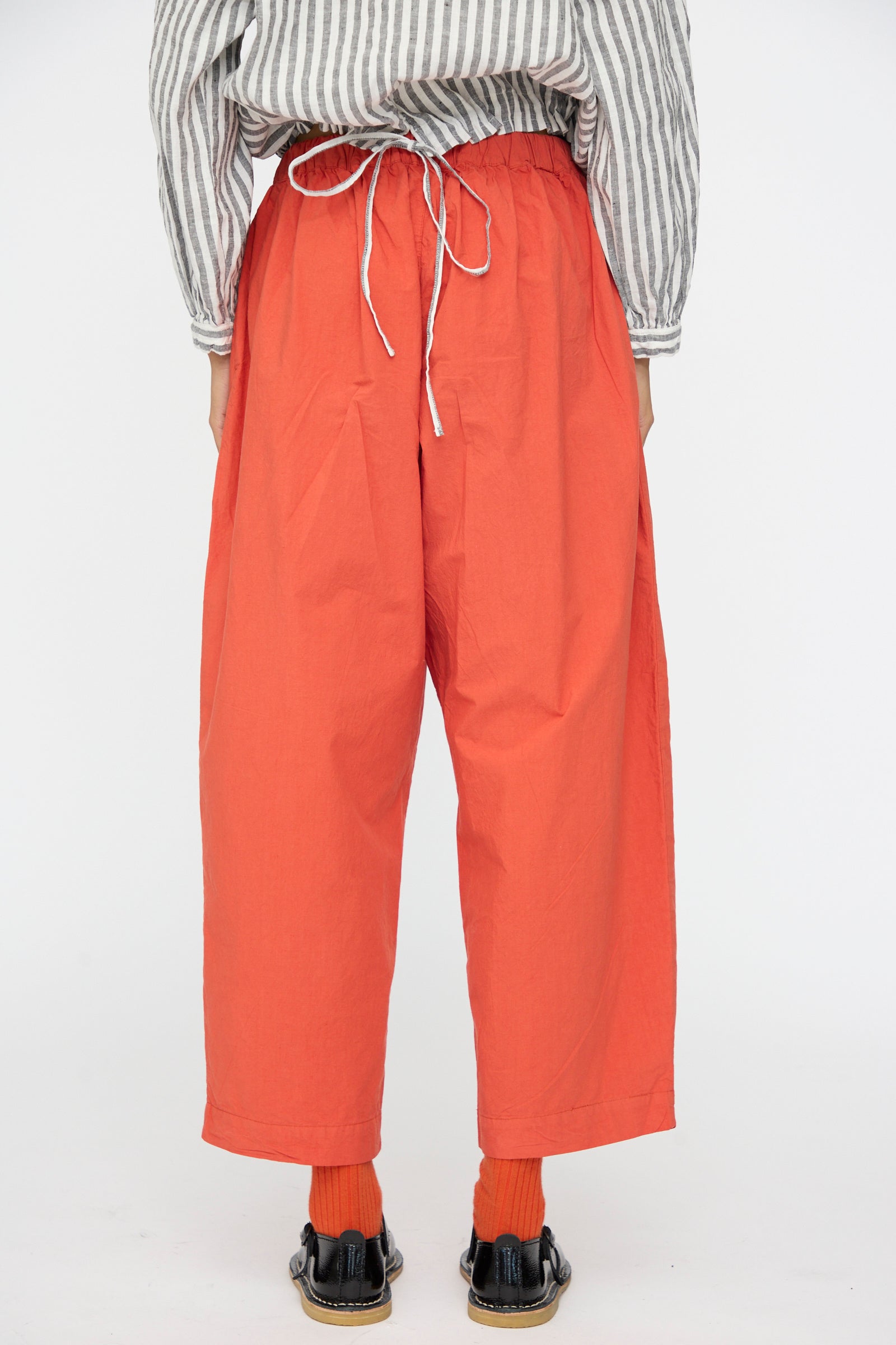 Person wearing nest Robe Cotton Linen Typewriter Omi-Zarashi Easy Pants in Orange, a striped long-sleeve shirt, red socks, and black shoes, photographed from the back against a plain white background.