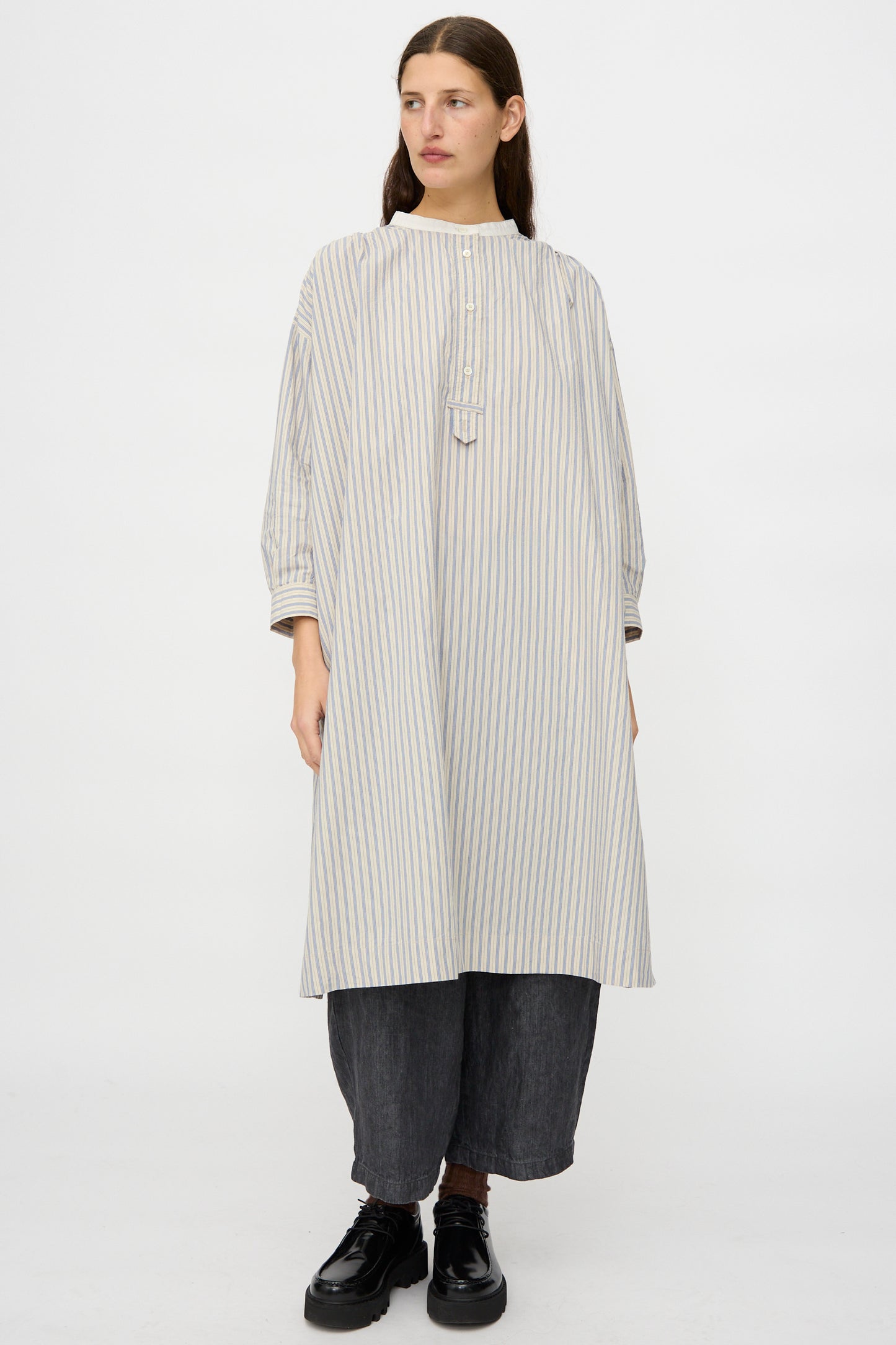 A person wearing the nest Robe Cotton Pull-on Dress in Stripe over dark wide-leg pants and black shoes, standing against a plain white background.