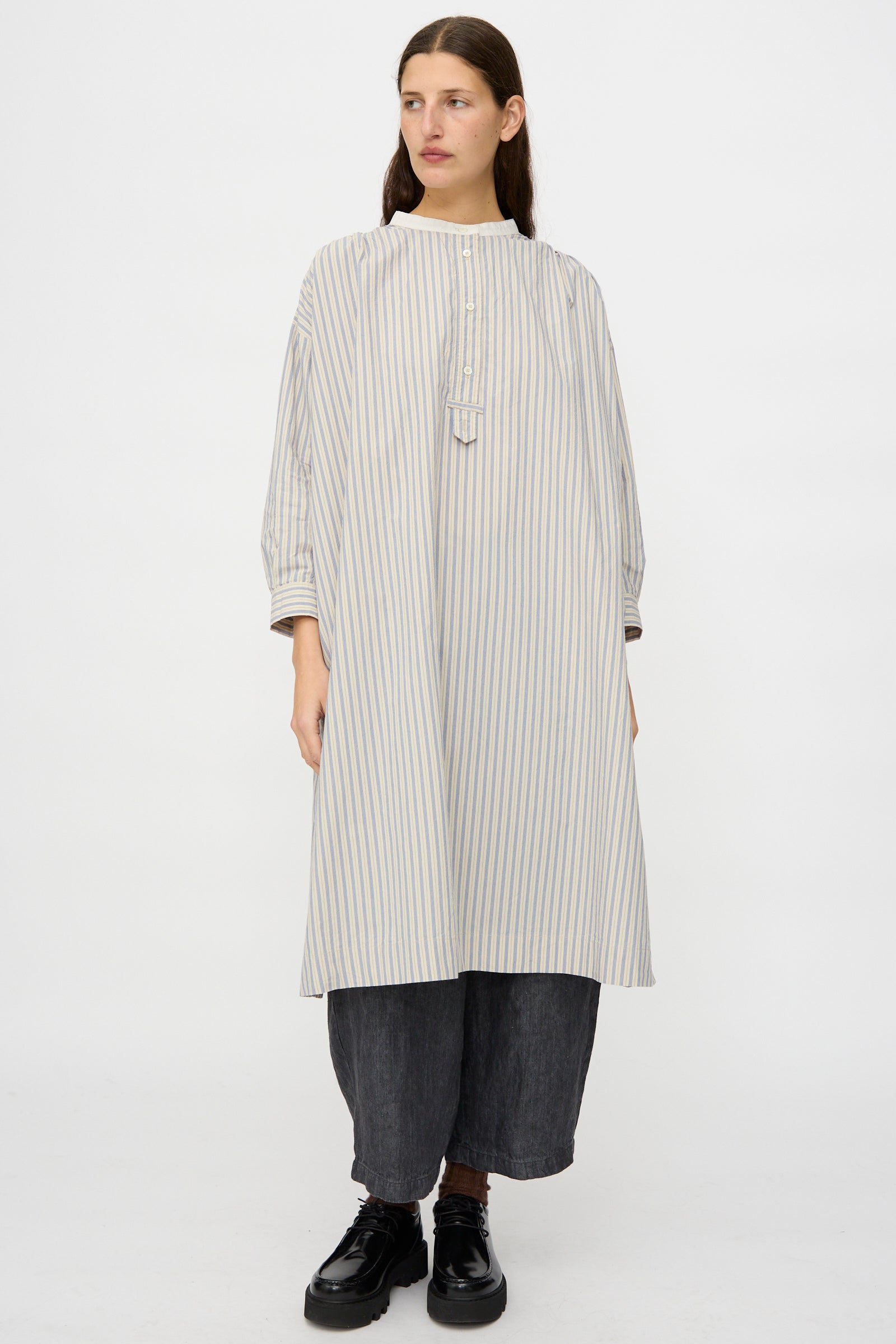 A person wearing the nest Robe Cotton Pull-on Dress in Stripe over dark wide-leg pants and black shoes, standing against a plain white background.