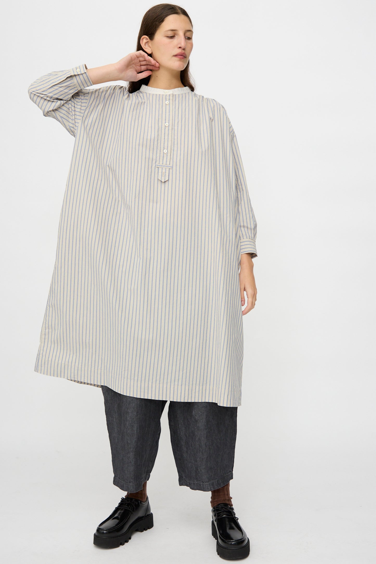 A person wears the nest Robe Cotton Pull-on Dress in Stripe over dark wide-leg pants and black shoes, standing against a plain white background.