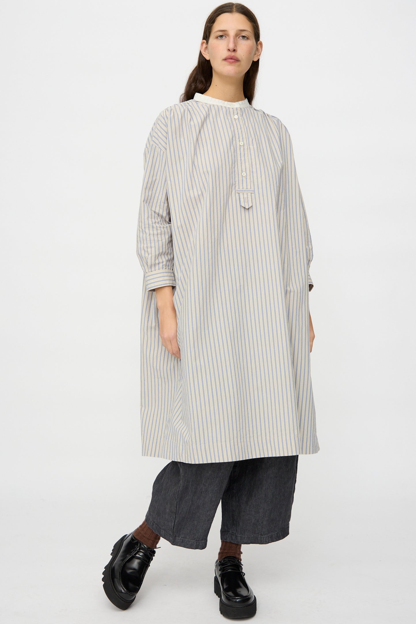 A person faces forward wearing the nest Robe Cotton Pull-on Dress in Stripe styled loosely over wide-leg dark pants and black shoes, set against a plain white background.