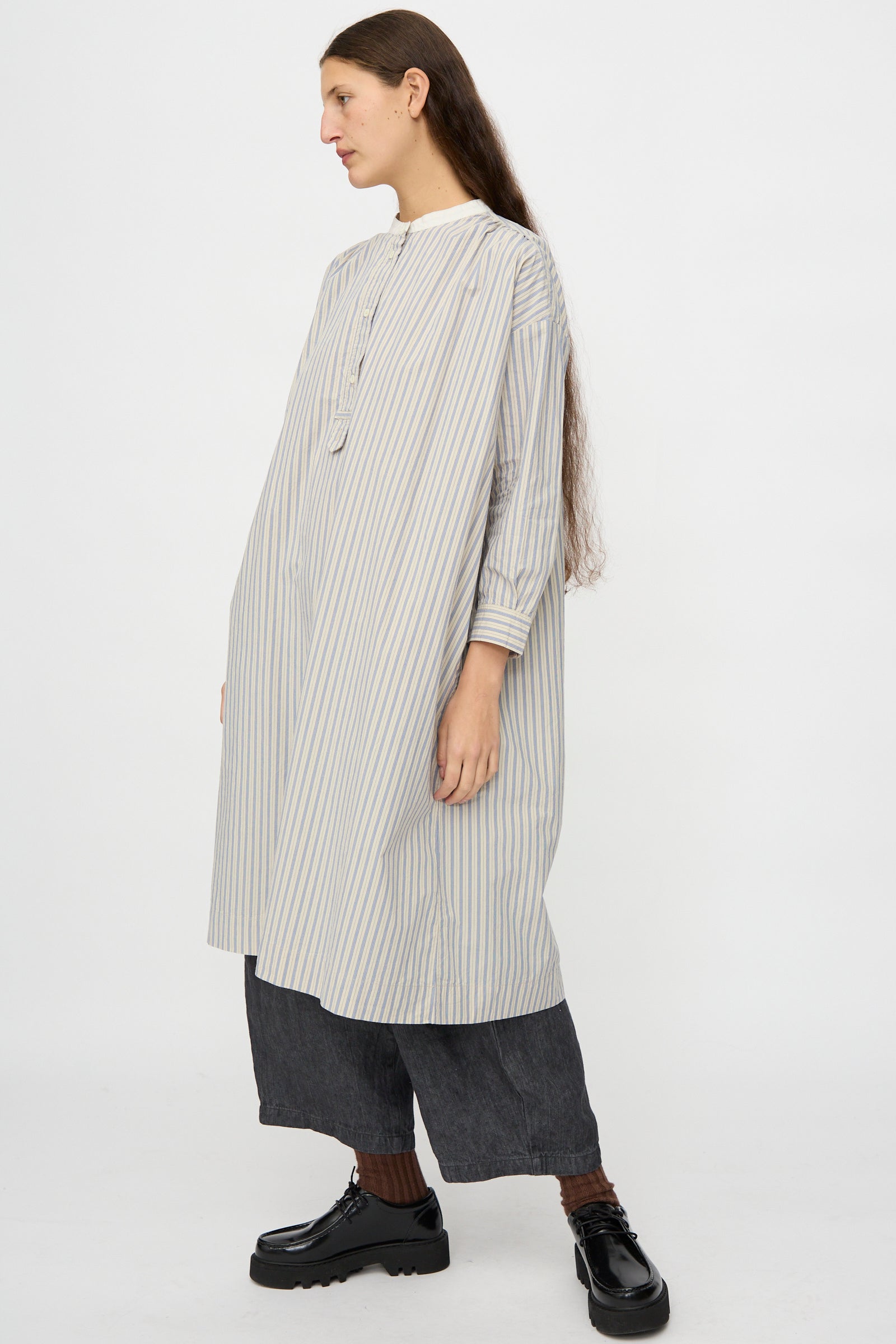 A person stands in profile against a white background wearing the nest Robe Cotton Pull-on Dress in Stripe, paired with dark wide-leg pants and black platform shoes.