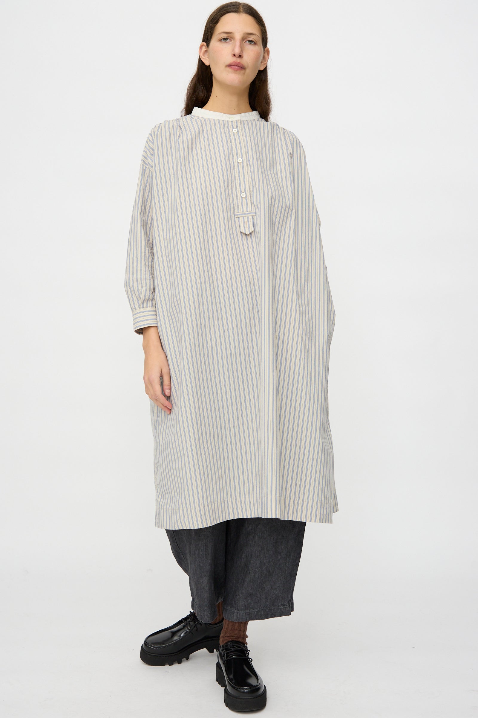 A person wears the nest Robe Cotton Pull-on Dress in Stripe over dark wide-leg pants and black shoes, standing against a plain background.