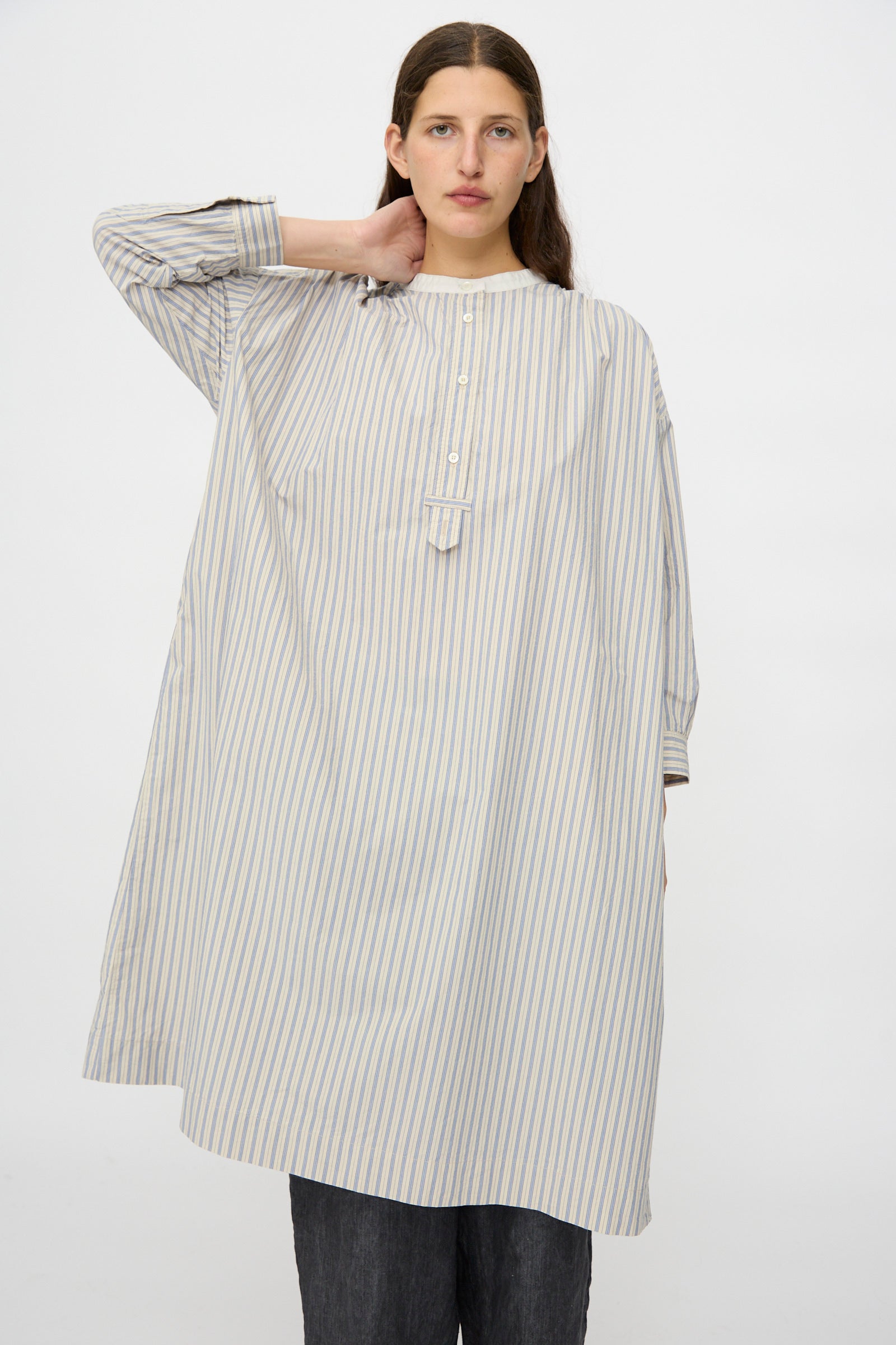 A person with long brown hair wears the nest Robe Cotton Pull-on Dress in Stripe, standing against a plain white background with one hand behind their neck.