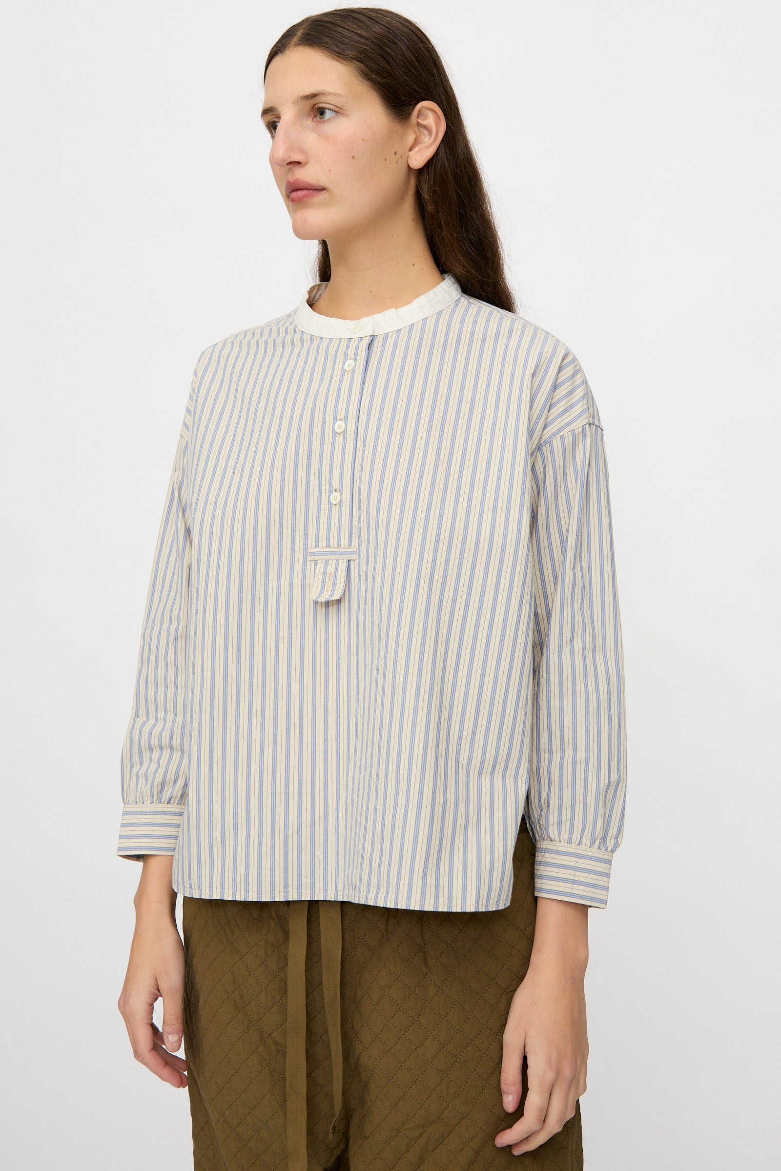 A person with long brown hair wears the nest Robe Cotton Pull-on Shirt in Stripe, featuring a relaxed fit and long sleeves, paired with olive green quilted pants, standing against a plain white background.