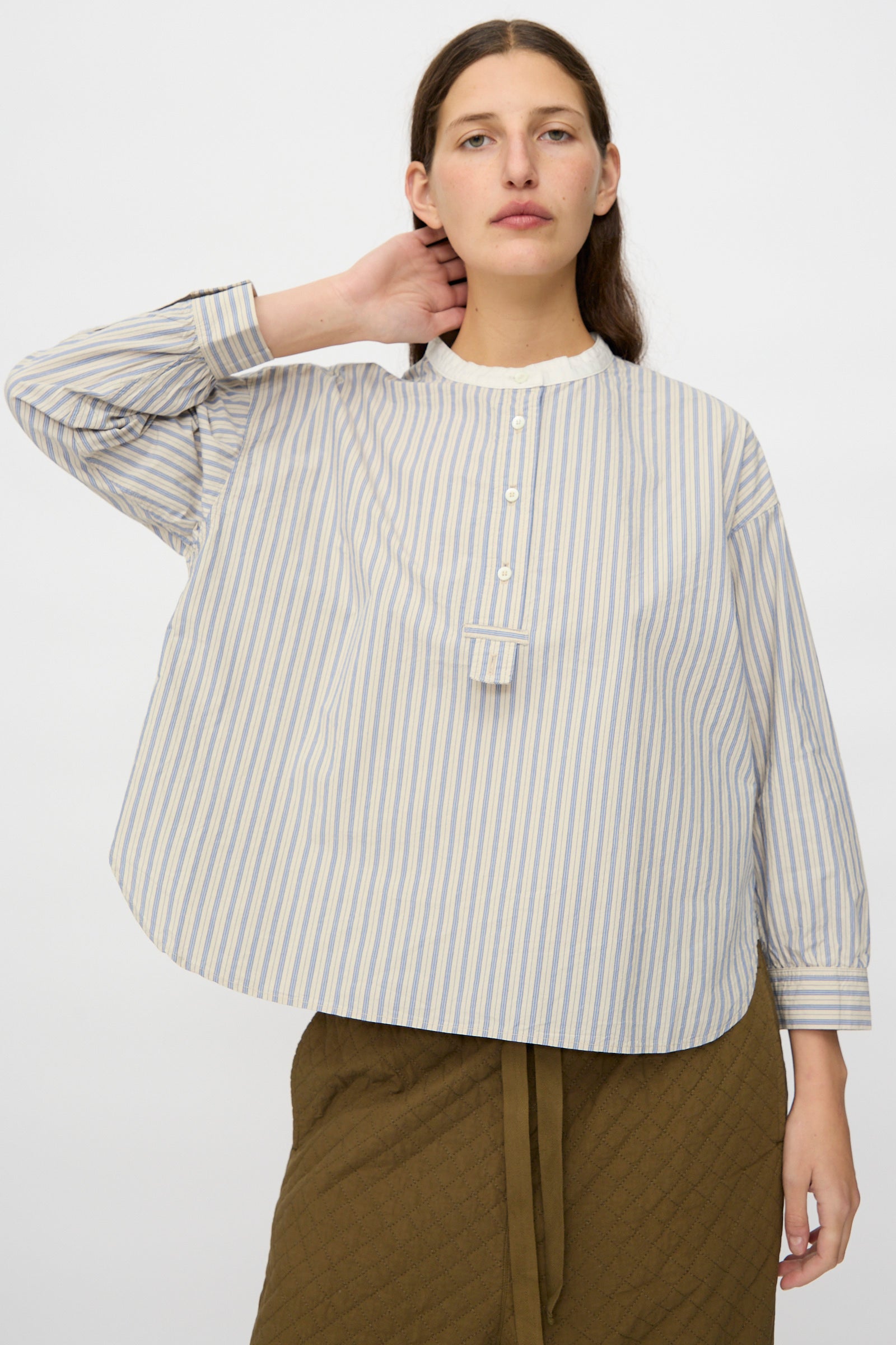 A person wearing the nest Robe Cotton Pull-on Shirt in Stripe, featuring light blue and white stripes and a band collar with buttons, paired with an olive green skirt and posing with one hand behind their neck.
