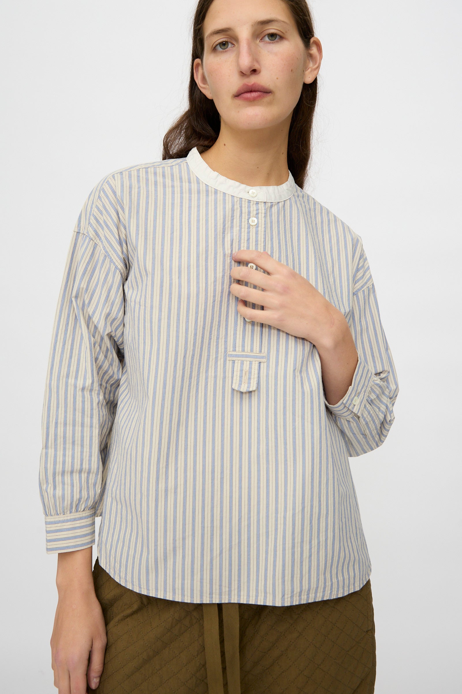 Wearing nest Robe's Cotton Pull-on Shirt in Stripe—a relaxed fit, blue and white striped cotton top with a white collar and button placket—paired with olive quilted pants, the person stands against a plain light background.