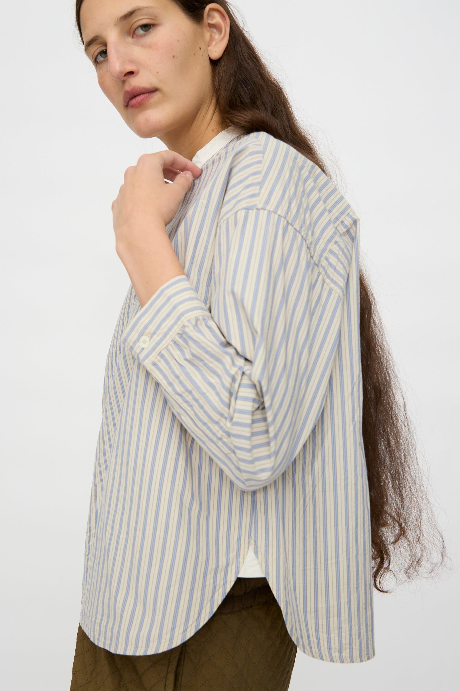 A person with long brown hair wears the nest Robe Cotton Pull-on Shirt in Stripe and brown pants, standing sideways against a plain light background.