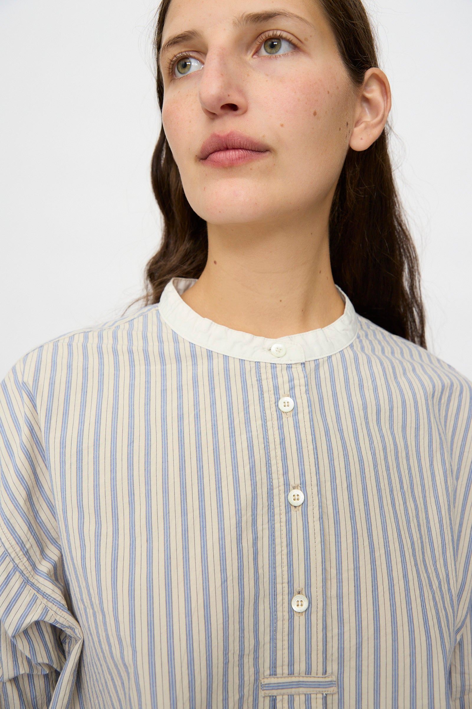 A person with long brown hair wears the nest Robe Cotton Pull-on Shirt in Stripe, featuring a relaxed fit with vertical blue stripes and white buttons, looking slightly off to the side against a plain background.