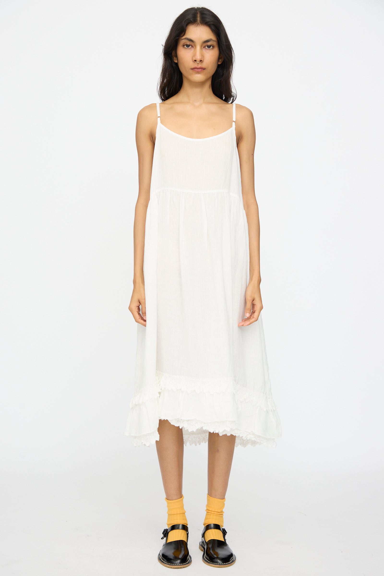 A woman stands facing forward against a white background wearing the nest Robe Linen Cotton Lace-Hem Cami Dress in Off White, styled with yellow socks and black sandals.