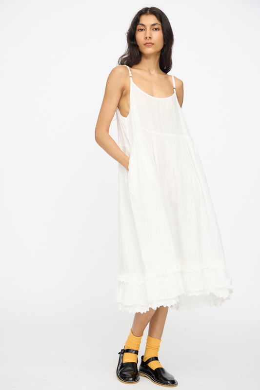 A person wears the nest Robe Linen Cotton Lace-Hem Cami Dress in Off White, paired with yellow socks and black shoes, standing against a plain white background.