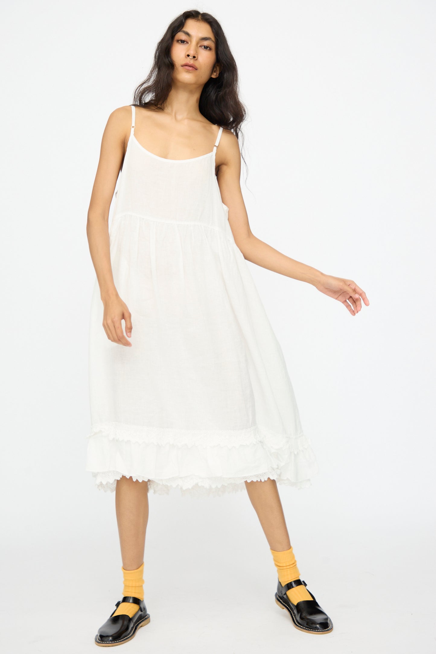 A person stands against a plain white background wearing the nest Robe Linen Cotton Lace-Hem Cami Dress in Off White, styled with yellow socks and black sandals.