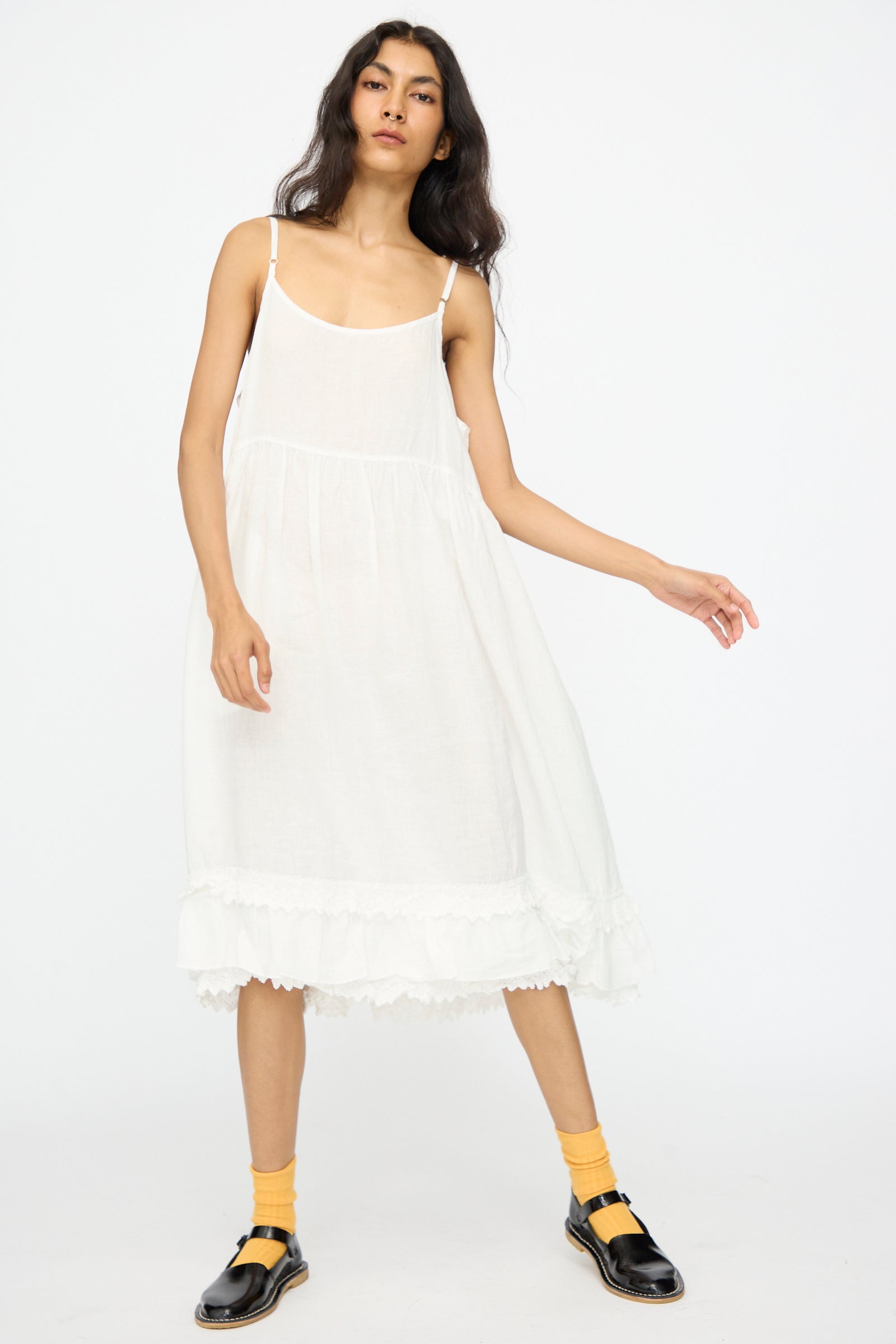 A person stands against a plain white background wearing the nest Robe Linen Cotton Lace-Hem Cami Dress in Off White, styled with yellow socks and black sandals.