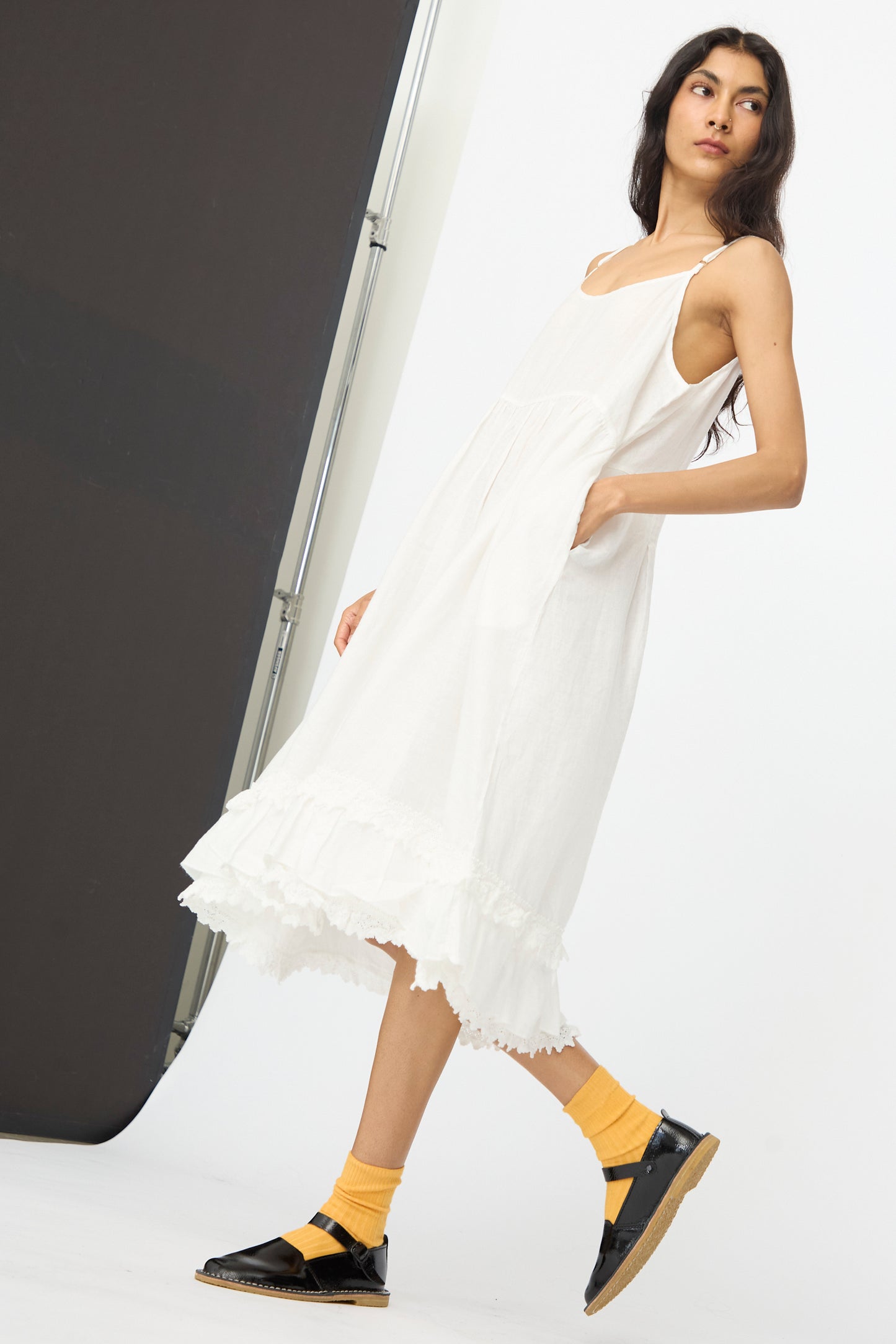 A woman models the nest Robe Linen Cotton Lace-Hem Cami Dress in Off White, paired with yellow socks and black sandals, standing against a simple studio backdrop.