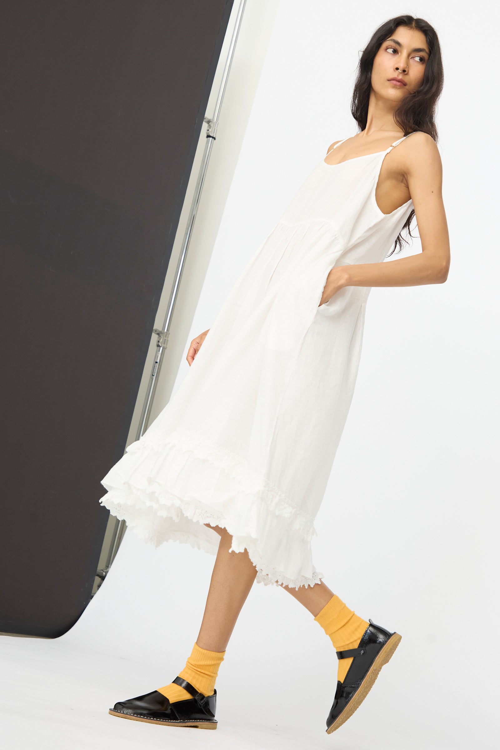 A woman models the nest Robe Linen Cotton Lace-Hem Cami Dress in Off White, paired with yellow socks and black sandals, standing against a simple studio backdrop.
