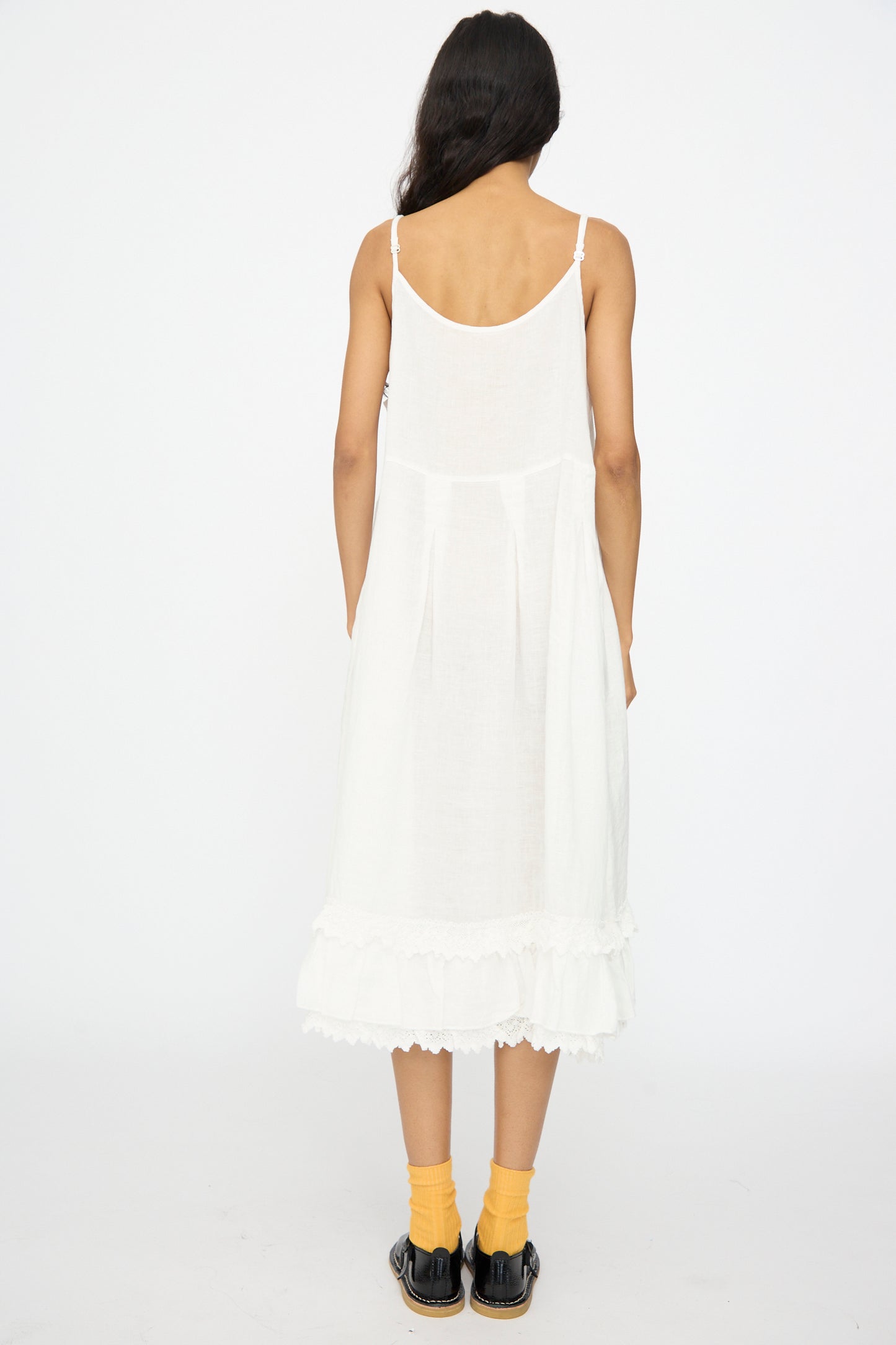 A person faces away from the camera wearing the nest Robe Linen Cotton Lace-Hem Cami Dress in Off White, paired with yellow socks and black shoes, set against a plain white background.