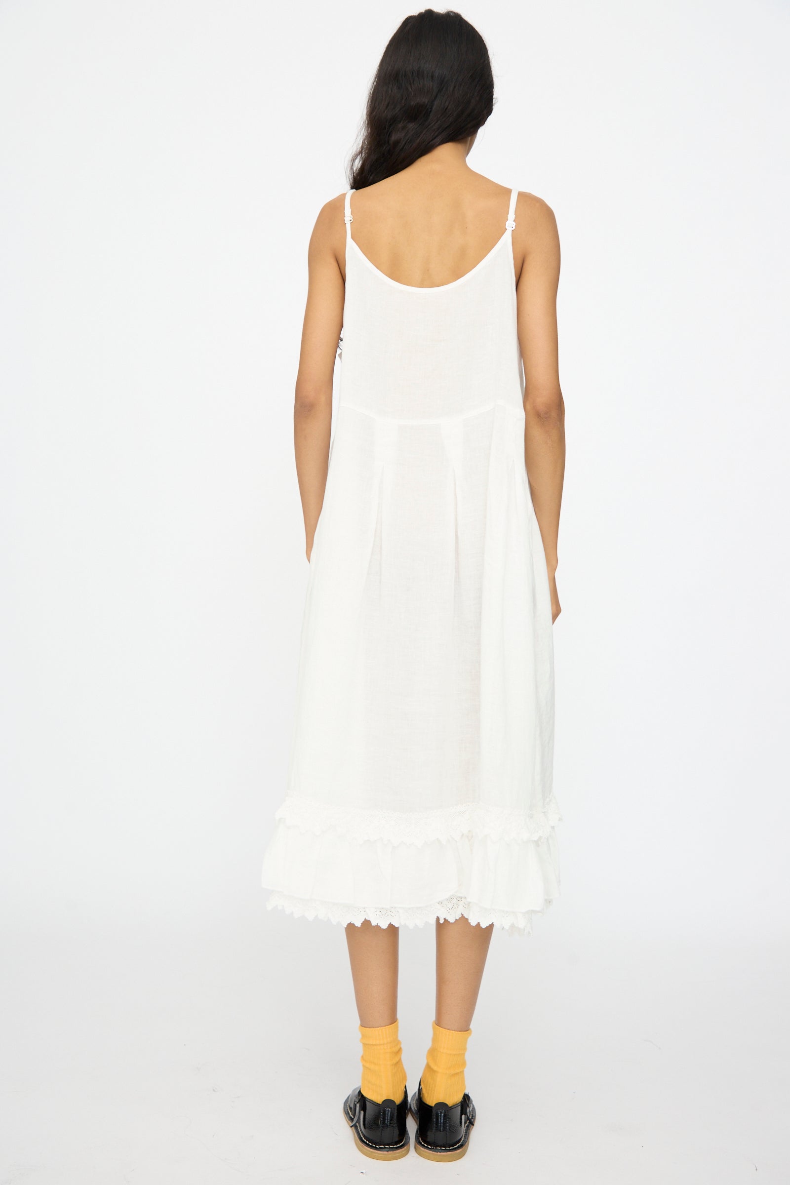 A person faces away from the camera wearing the nest Robe Linen Cotton Lace-Hem Cami Dress in Off White, paired with yellow socks and black shoes, set against a plain white background.