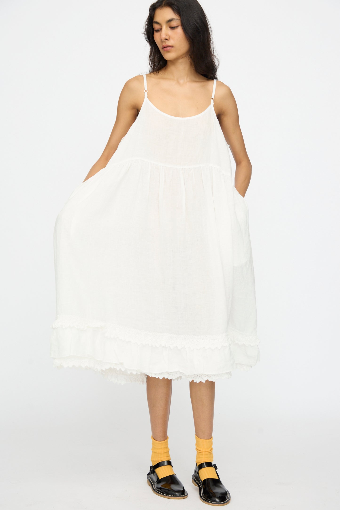 A person wears the nest Robe Linen Cotton Lace-Hem Cami Dress in Off White, yellow socks, and black shoes while standing against a plain white background.