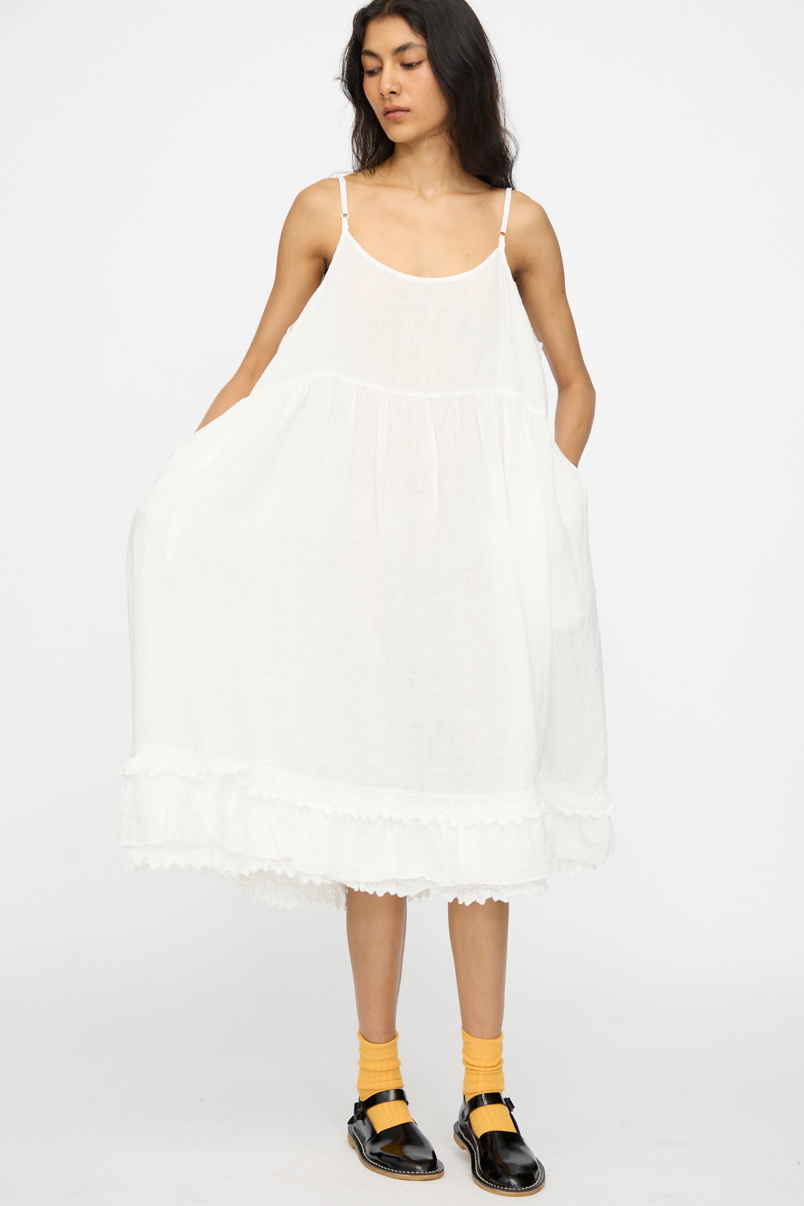 A person wears the nest Robe Linen Cotton Lace-Hem Cami Dress in Off White, yellow socks, and black shoes while standing against a plain white background.