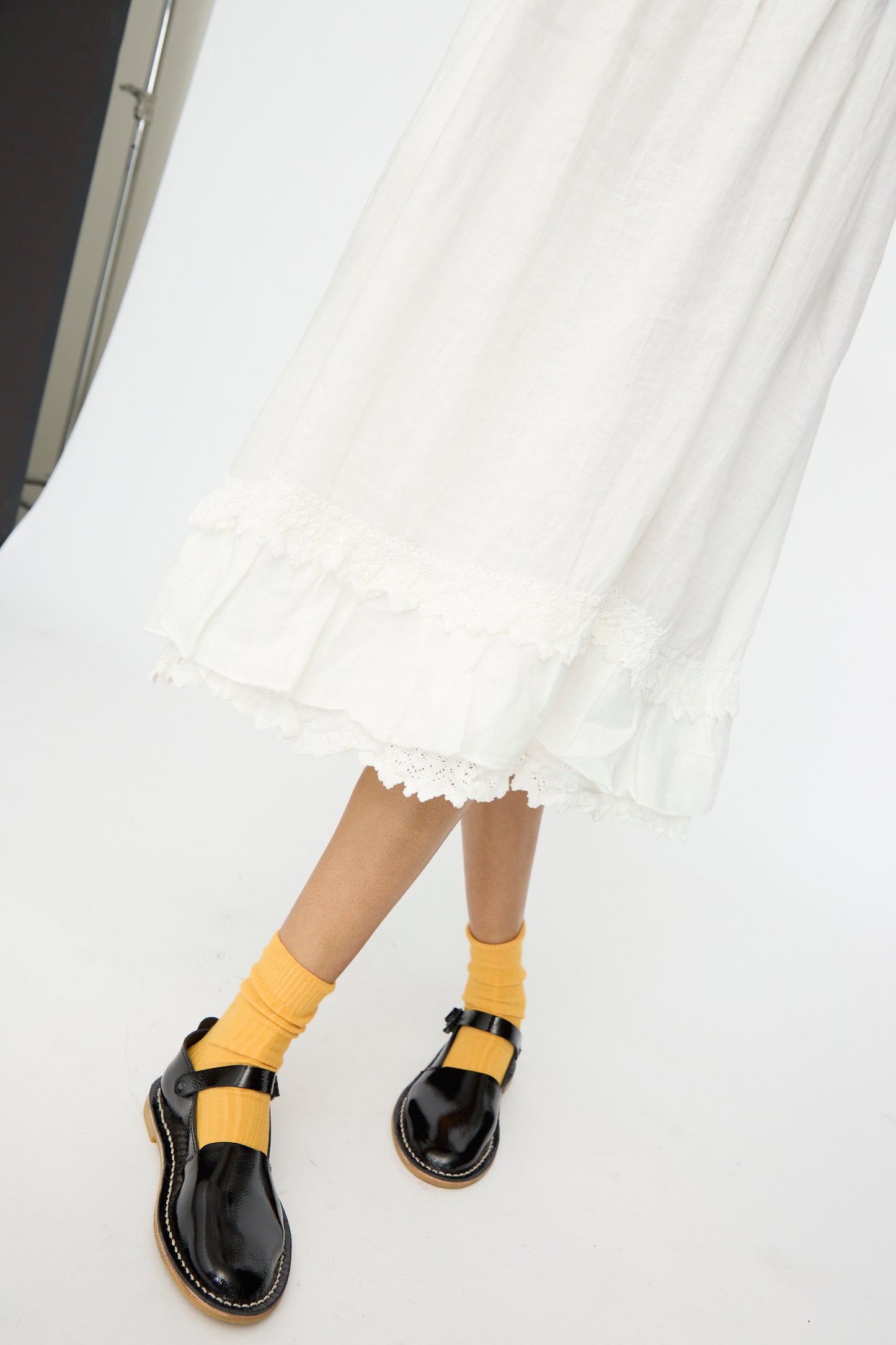 A person wearing the nest Robe Linen Cotton Lace-Hem Cami Dress in Off White, yellow socks, and black patent shoes stands on a white background.