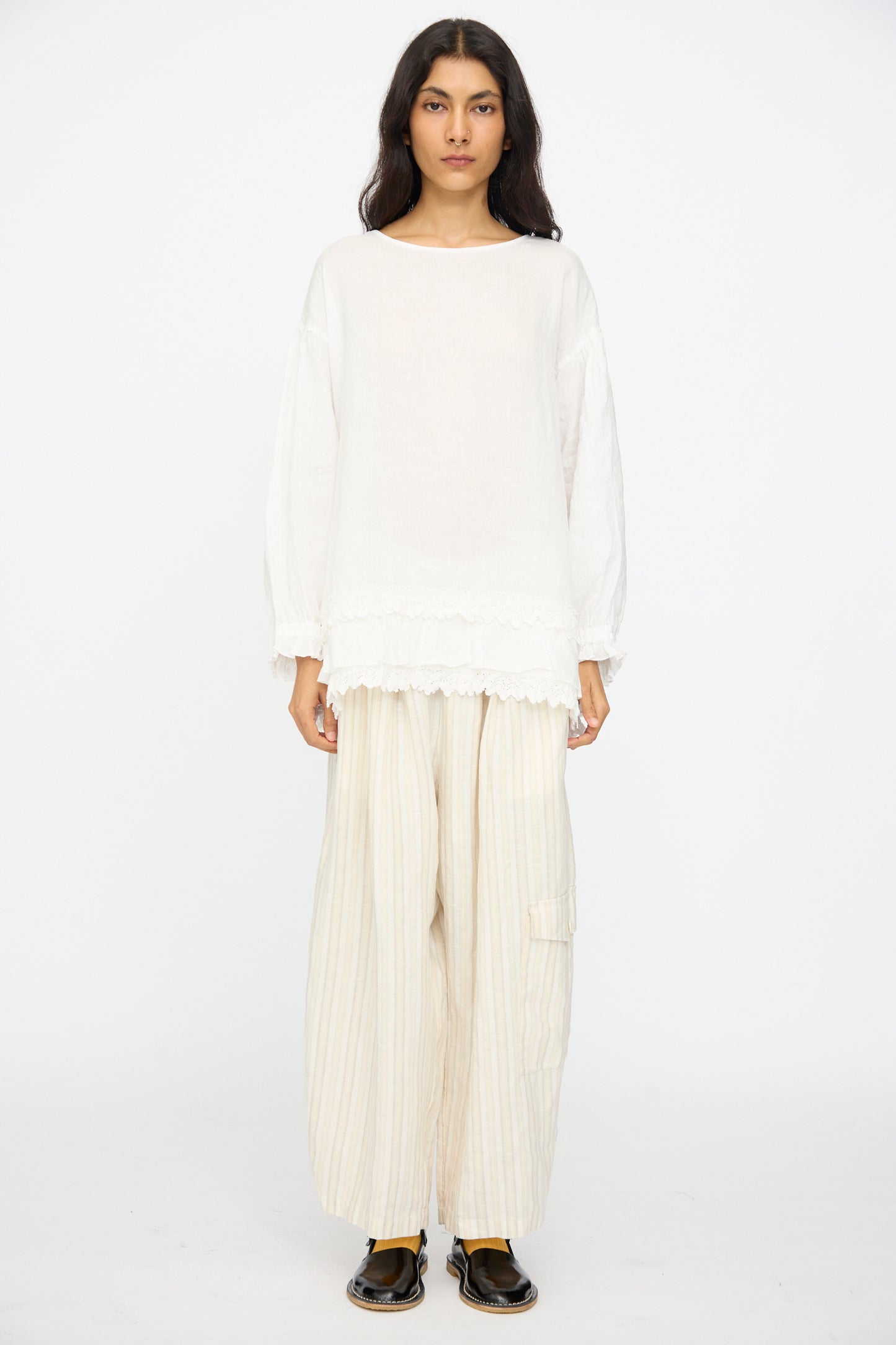 A woman stands facing forward against a plain white background, wearing the nest Robe Linen Cotton Lace-Hem Two Way Blouse in Off White, paired with wide-leg, light-striped pants and black shoes.